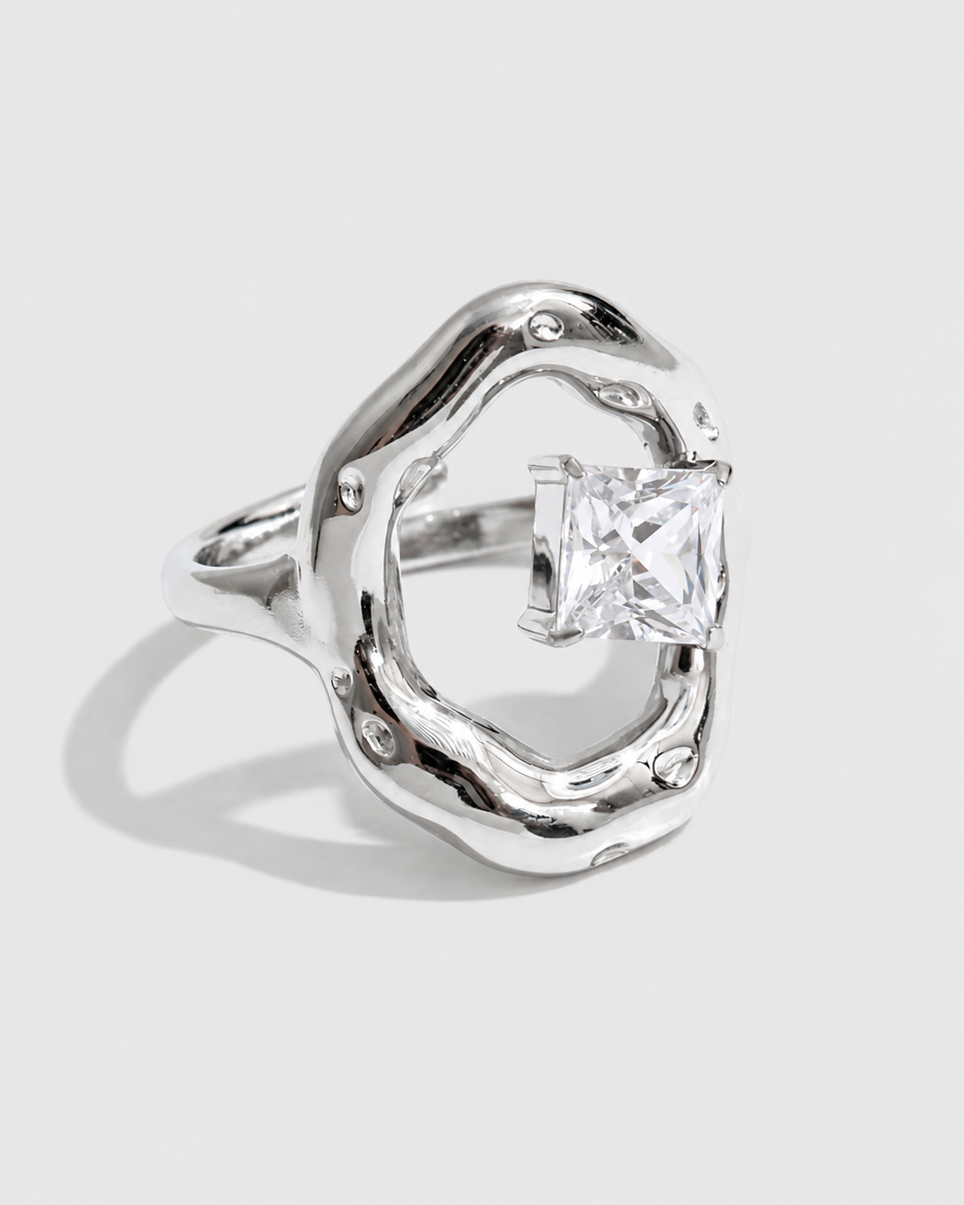 A silver ring with an organic, wavy openwork design and a central princess-cut clear gemstone, shown on a light gray background.