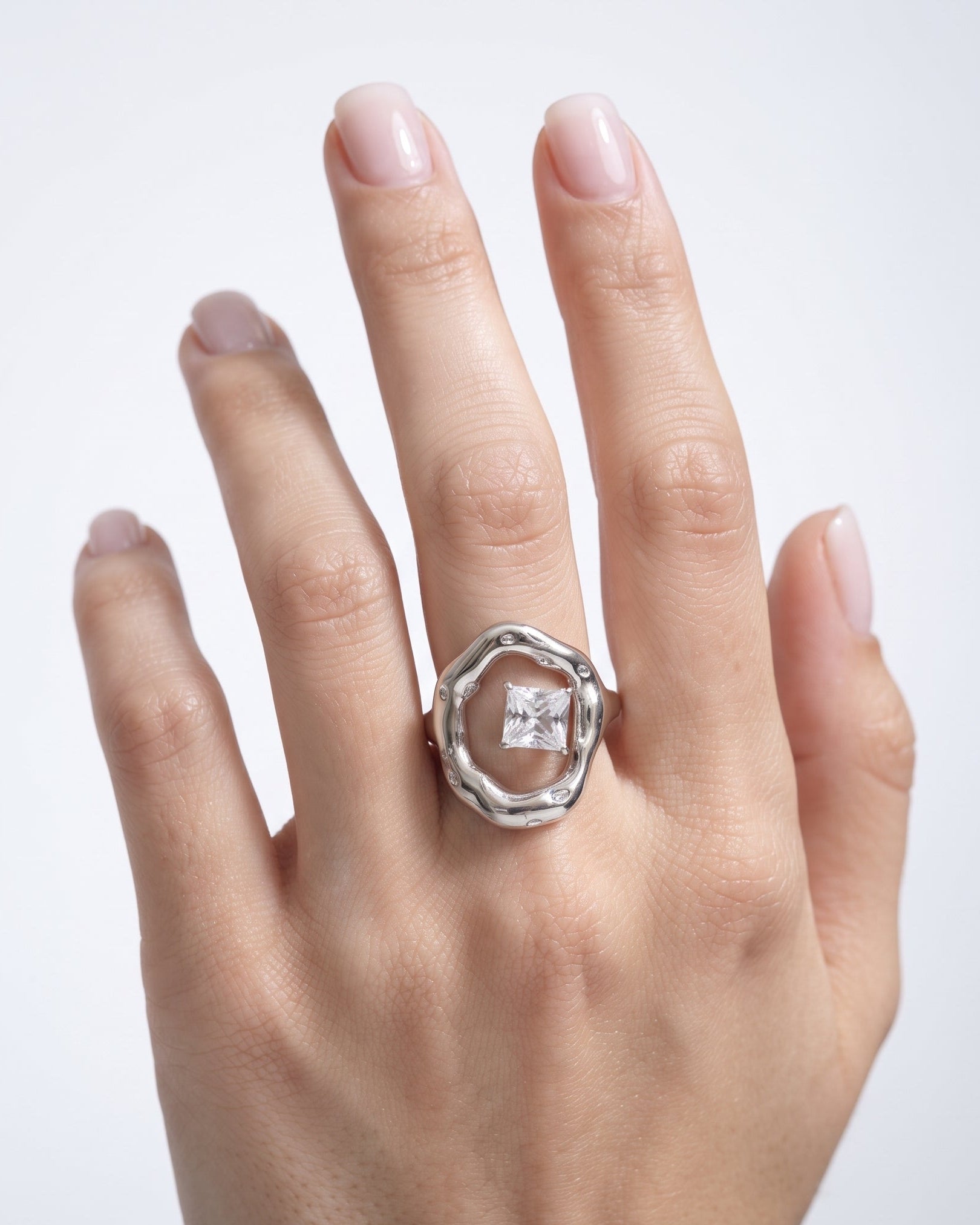 A silver ring with a square-cut clear gemstone set in a textured, organic-shaped bezel, shown worn on a hand against a light gray background.