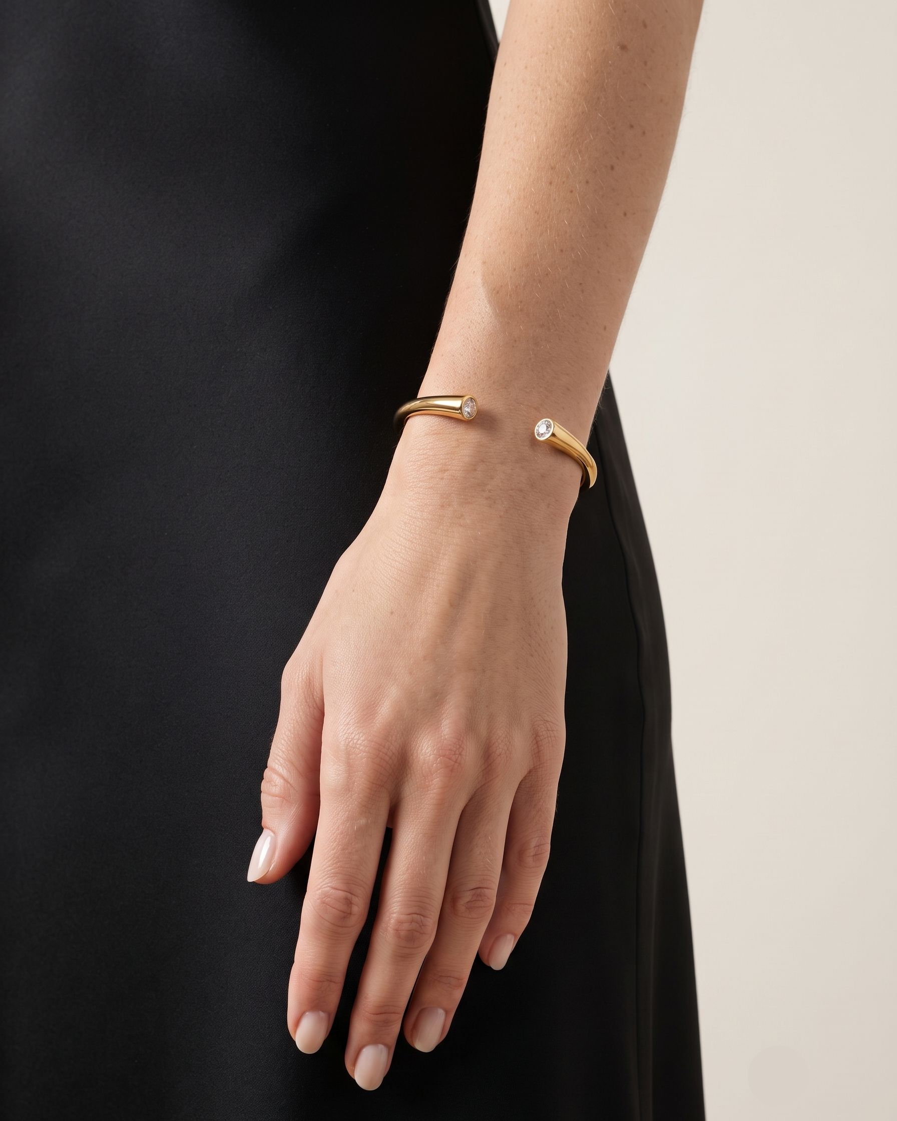 A close-up of a person's hand and wrist wearing a slim gold cuff bracelet with a small clear gemstone on one end, paired with a black dress against a light neutral background.
