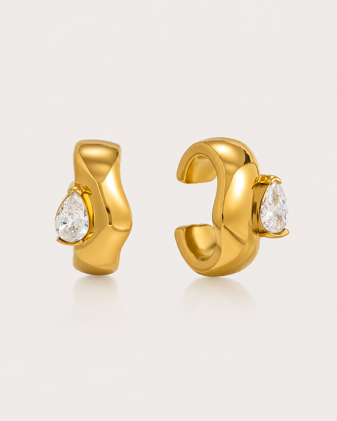 A pair of gold huggie hoop earrings with a polished, organic wavy design and a clear teardrop gemstone on each earring, set against a light beige background.
