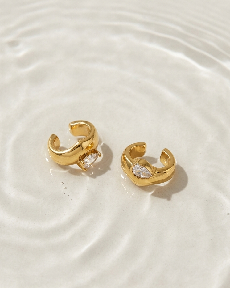 A pair of gold huggie hoop earrings with clear heart-shaped crystals, resting on a white surface with soft water ripples.