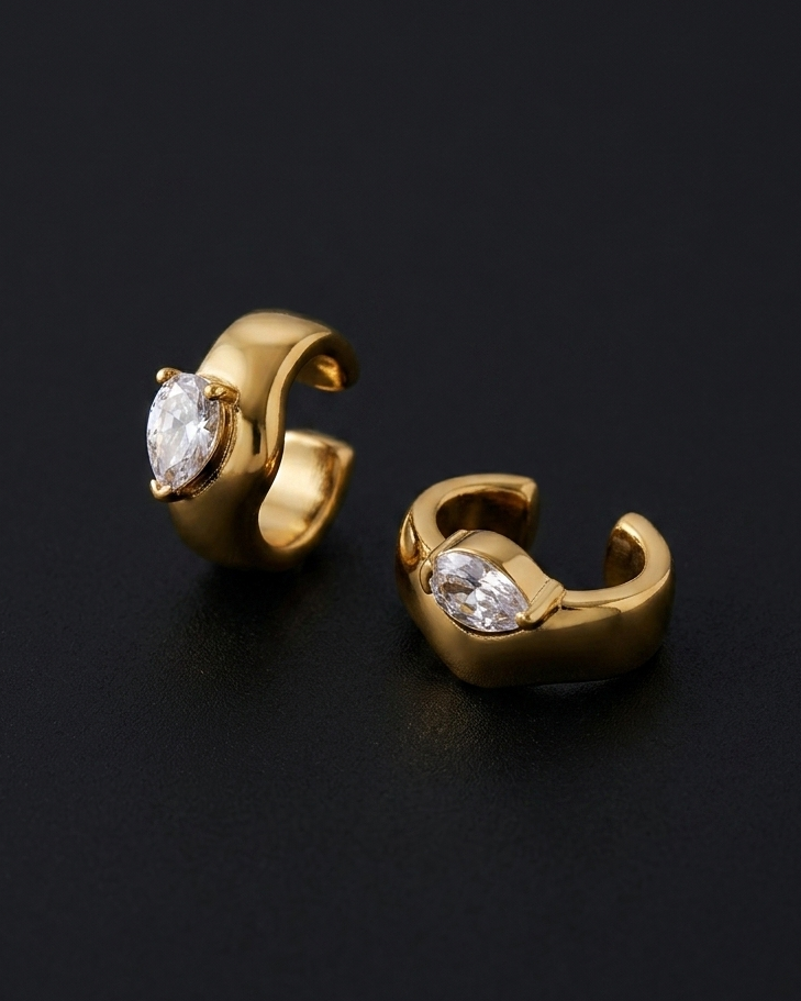 A pair of gold huggie hoop earrings with marquise-cut clear gemstones, displayed on a dark black background.