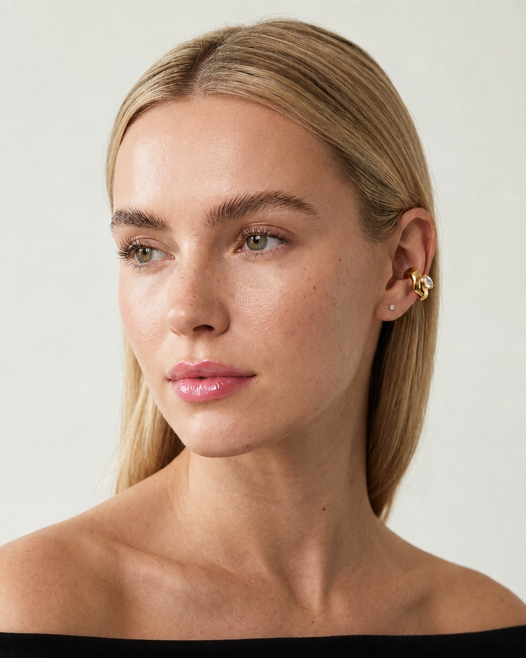 A close-up portrait of a blonde woman wearing a small gold hoop earring with a dangling textured gold charm, set against a plain light gray background.