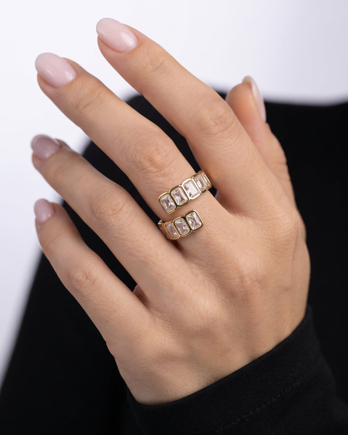 A close-up of a hand wearing two gold rings with rectangular clear stones set in a row, against a black and white background.