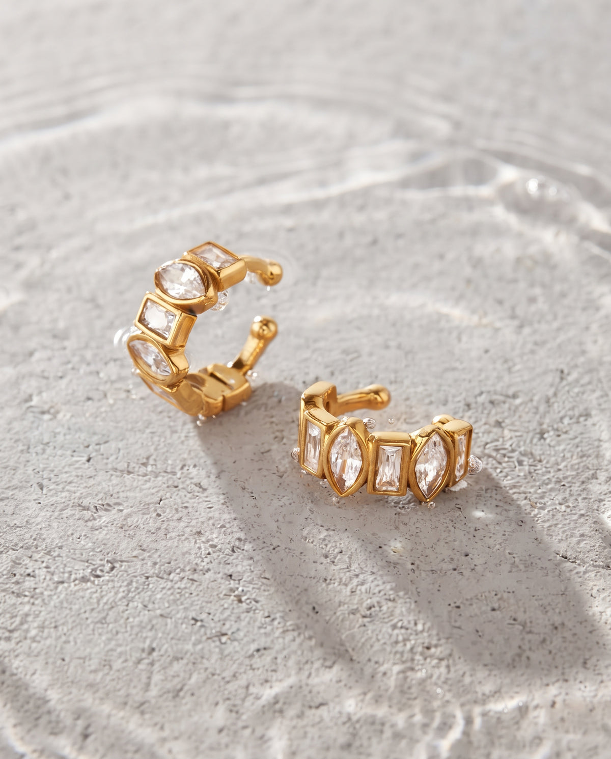 A pair of gold huggie hoop earrings with clear marquise and baguette gemstones, displayed on a textured light gray stone surface.