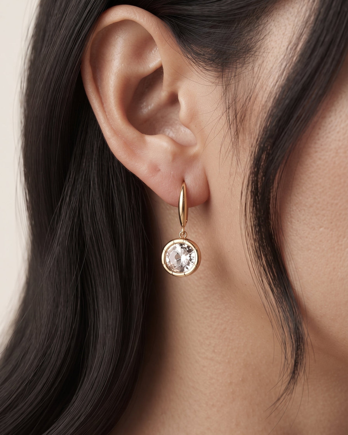 A close-up of a model's ear wearing a delicate gold huggie hoop earring with a round bezel-set clear gemstone.