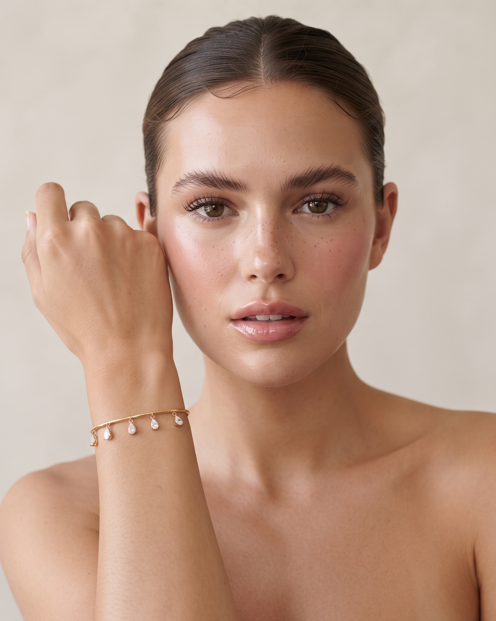 A close-up portrait of a woman wearing a delicate gold chain bracelet with small dangling pearl-like beads on her wrist, posed against a soft beige background.