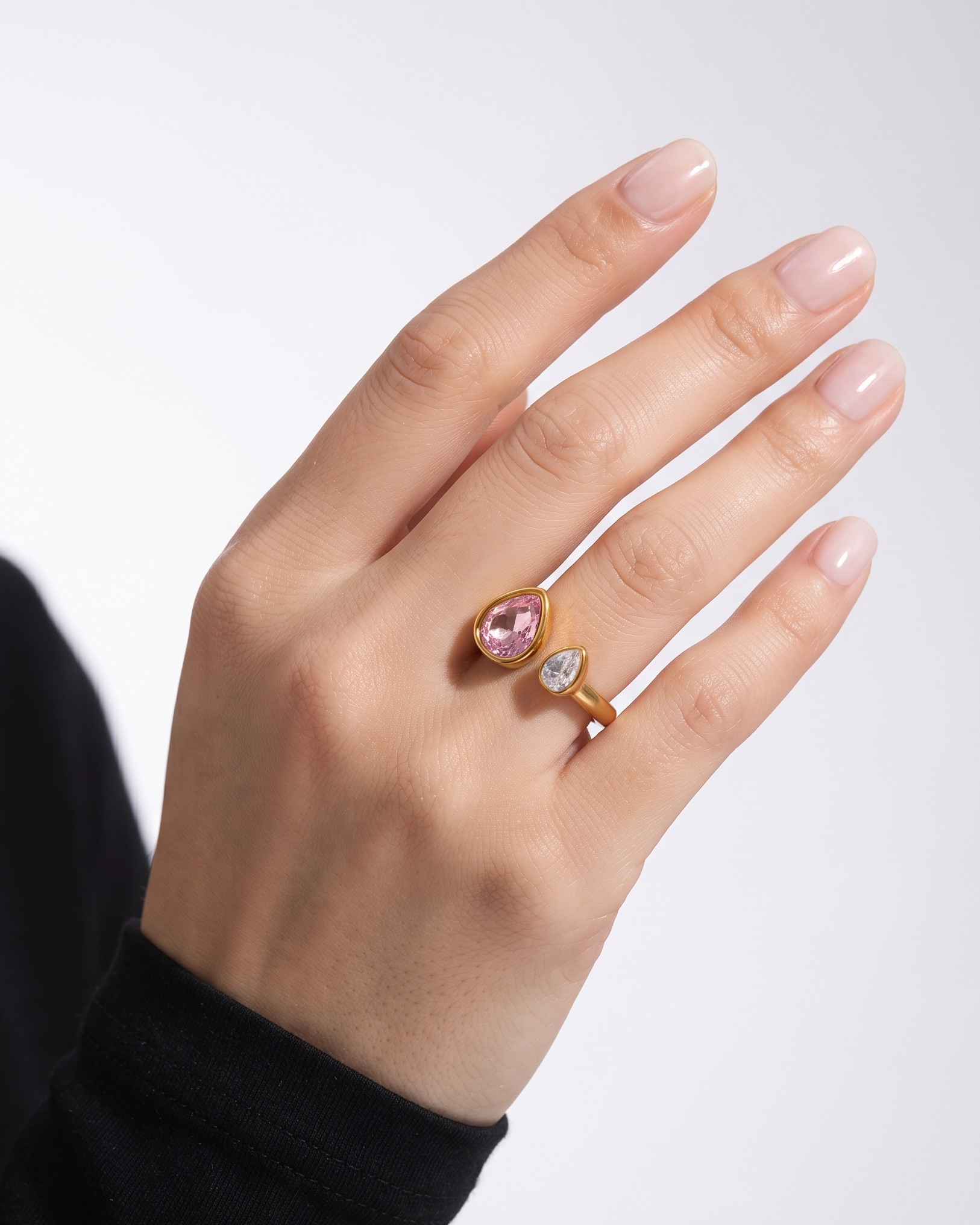 A close-up of a hand wearing two gold rings with teardrop-shaped pink and clear gemstones, set against a clean light gray background.