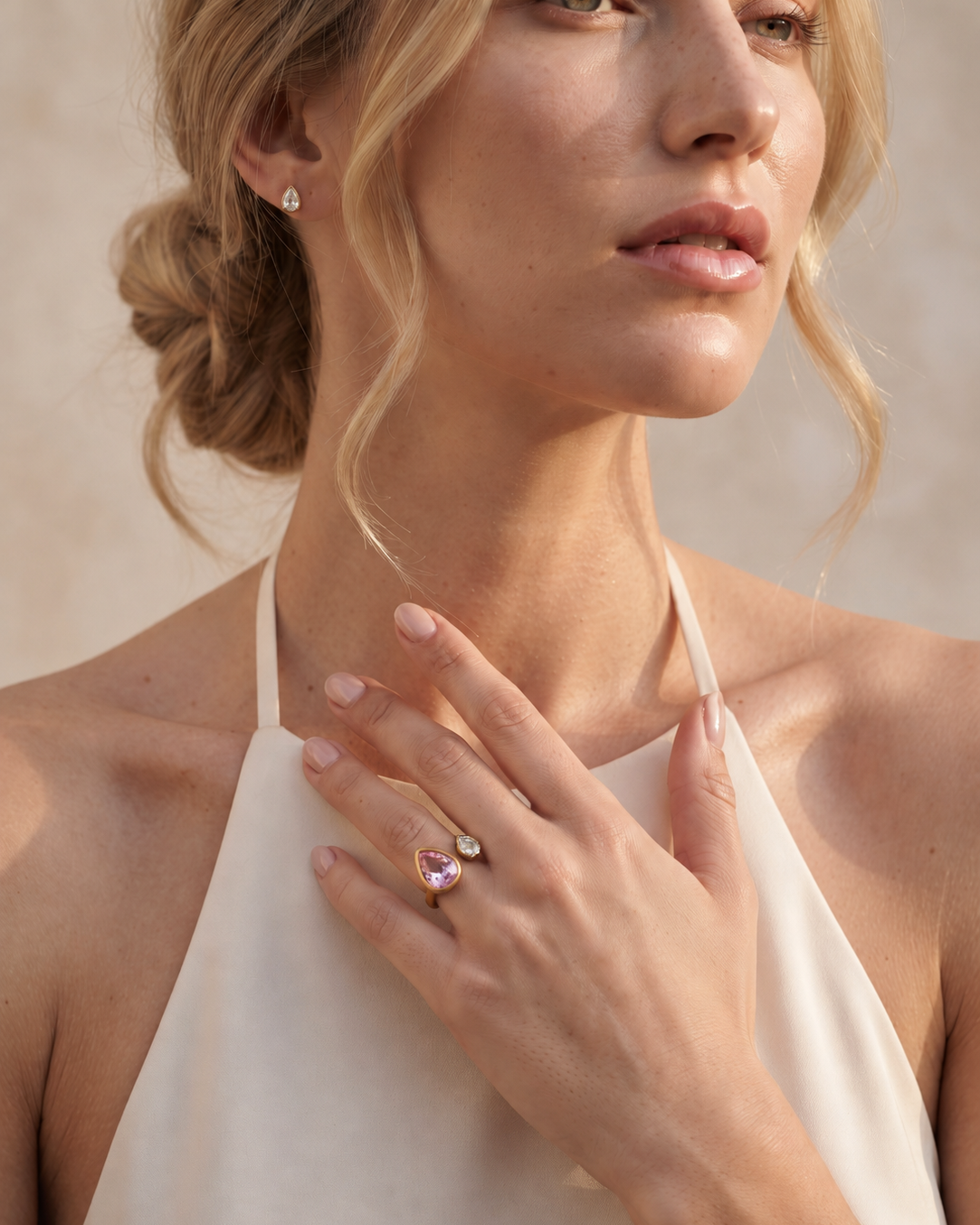 A close-up lifestyle shot of a woman wearing a delicate silver ring with a marquise-cut pink gemstone and a small clear accent stone on her finger, paired with a cream halter top against a warm beige background.