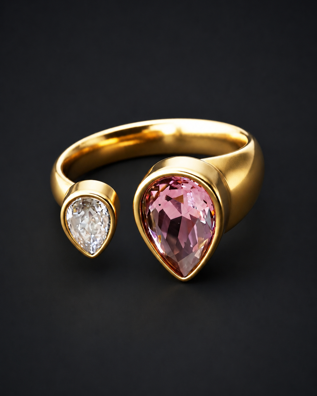 A gold open ring featuring a large faceted pink pear-cut gemstone and a smaller clear pear-cut gemstone, set against a dark black background.