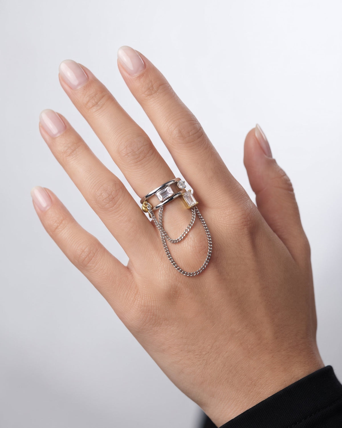 A close-up of a hand wearing a silver ring with a textured band, a small gold accent, and a dangling chain detail, set against a plain light gray background.