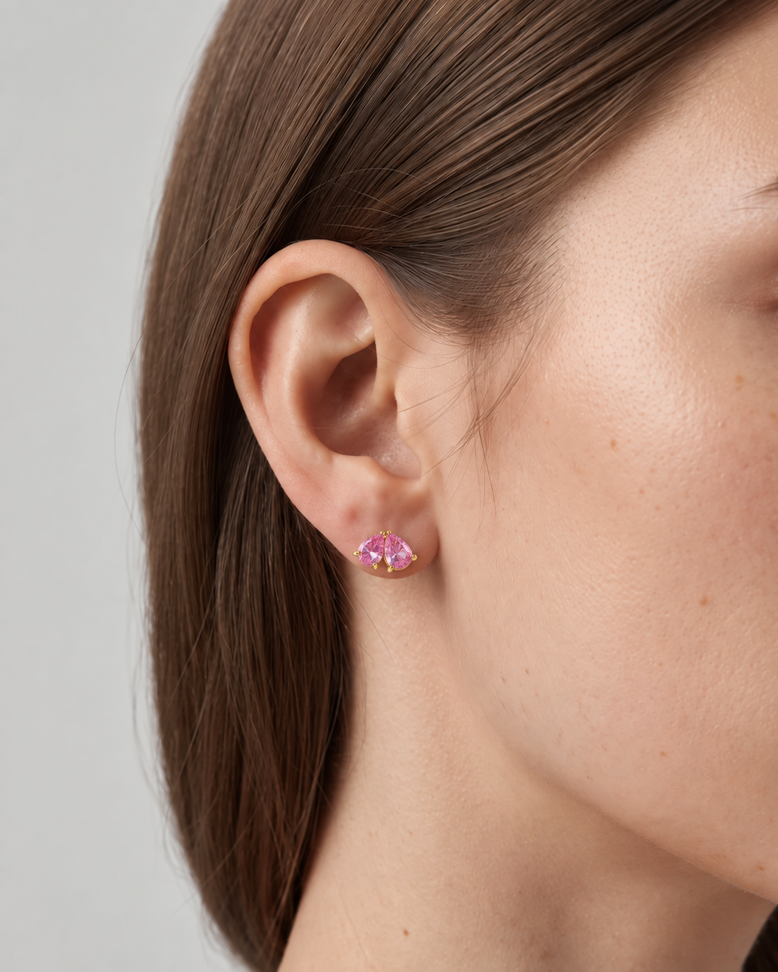 A close-up side profile of a woman's ear wearing a small gold stud earring with a pink marquise-cut gemstone.
