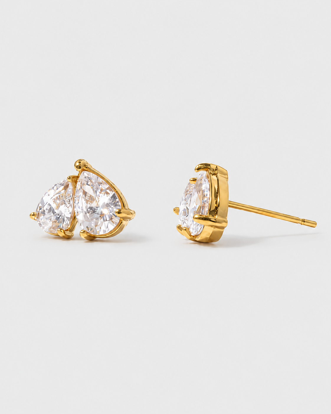 A pair of gold stud earrings with pear-cut clear gemstones in a prong setting, shown on a light gray background.
