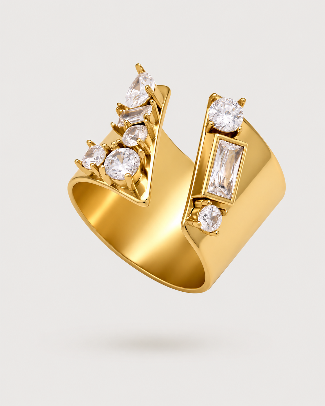 A gold statement ring featuring a wide, angular band adorned with multiple clear gemstones, including round, oval, and baguette cuts, set against a light gray background.