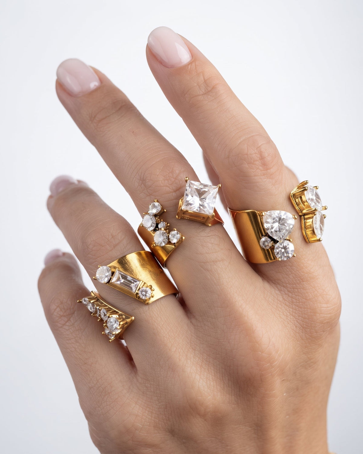 A close-up of a hand wearing multiple gold rings with clear gemstones, set against a clean light gray background.