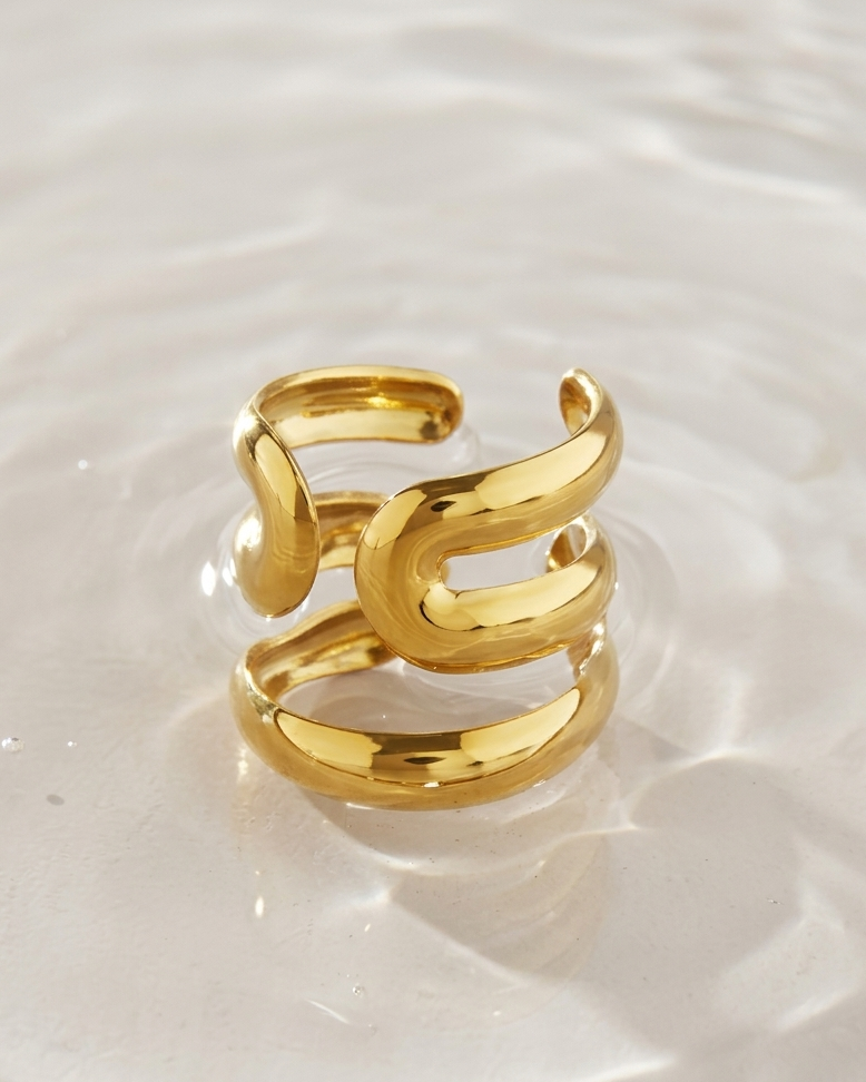 A polished gold open-band ring with a sculptural, layered wavy design, resting on a reflective surface of clear water.