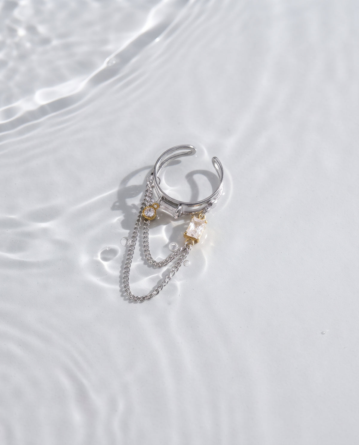 A silver-toned multi-layered ring with delicate chain drapes and small pearl-like and crystal accents, photographed on a white surface with soft water ripples.