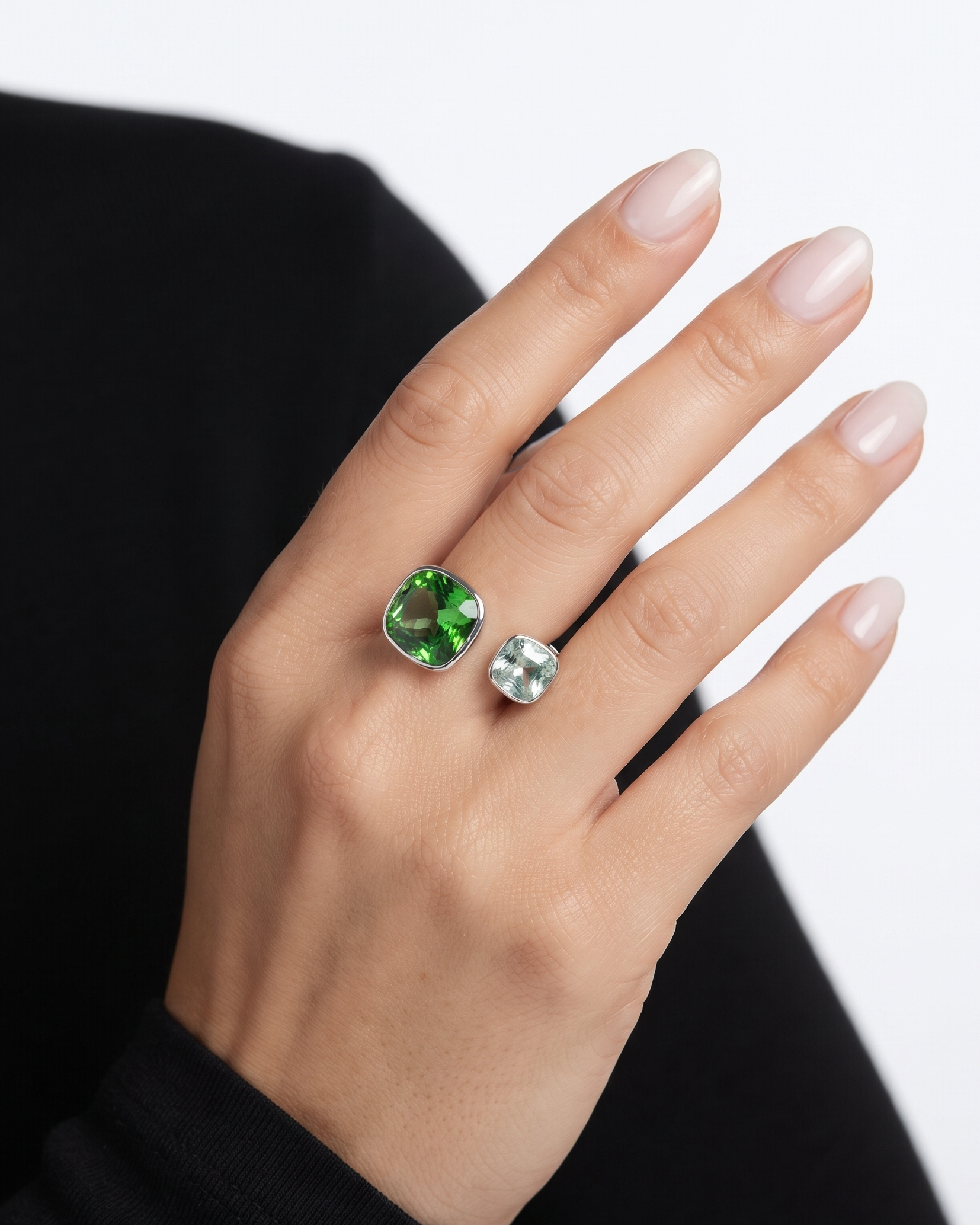 A close-up of a hand wearing two rings: a larger silver ring with a cushion-cut green gemstone and a smaller silver ring with a round clear gemstone, against a light gray background.