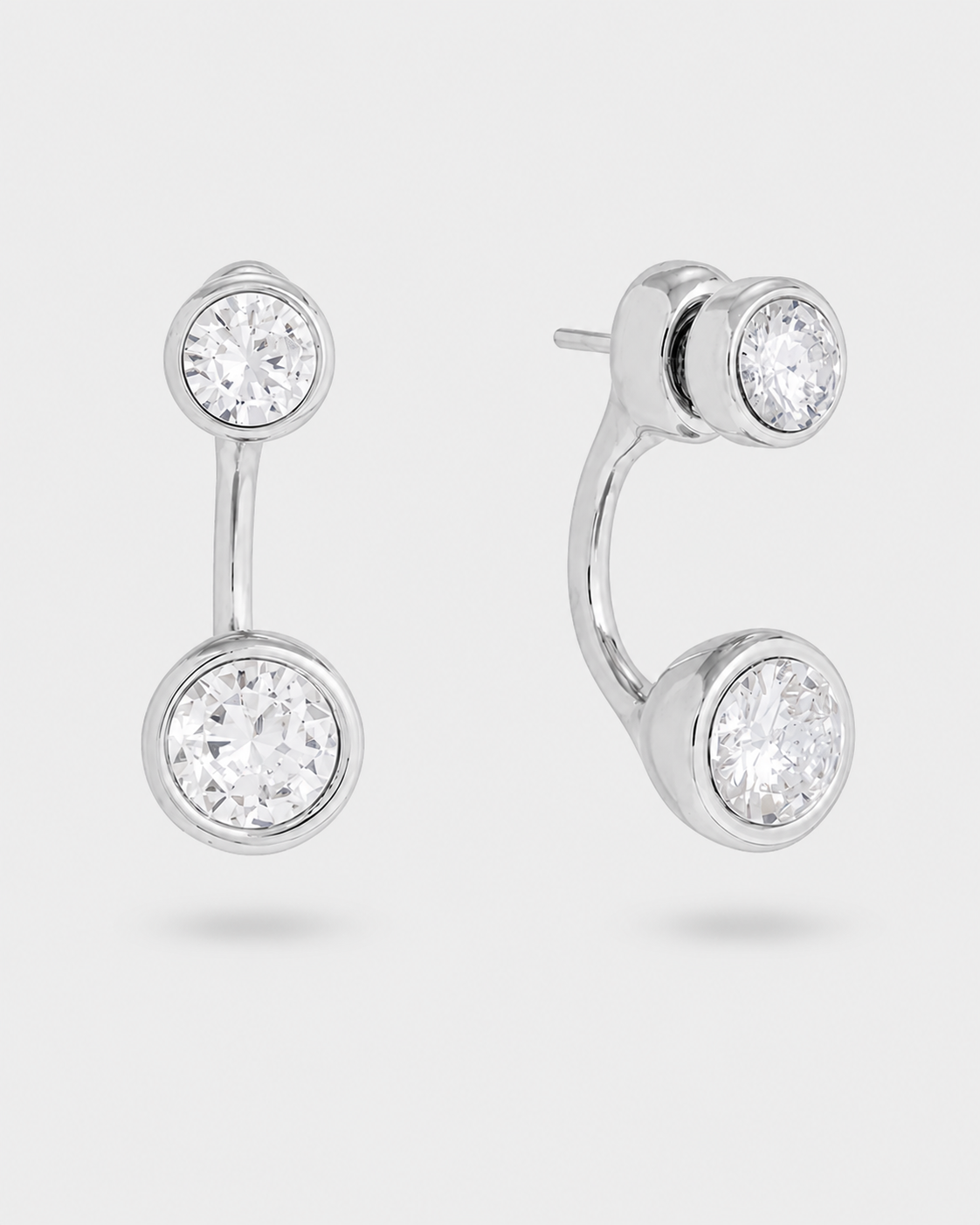 A pair of silver-toned stud earrings with round bezel-set clear gemstones, shown on a light gray background. One earring is shown front-facing and the other is angled to show the side profile and post.