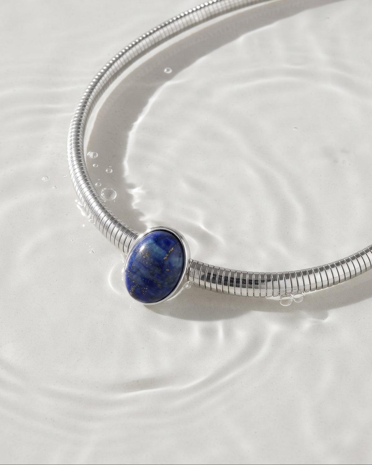 A silver snake-style bangle bracelet with an oval blue lapis lazuli stone centerpiece, resting on a surface of clear water with ripples.