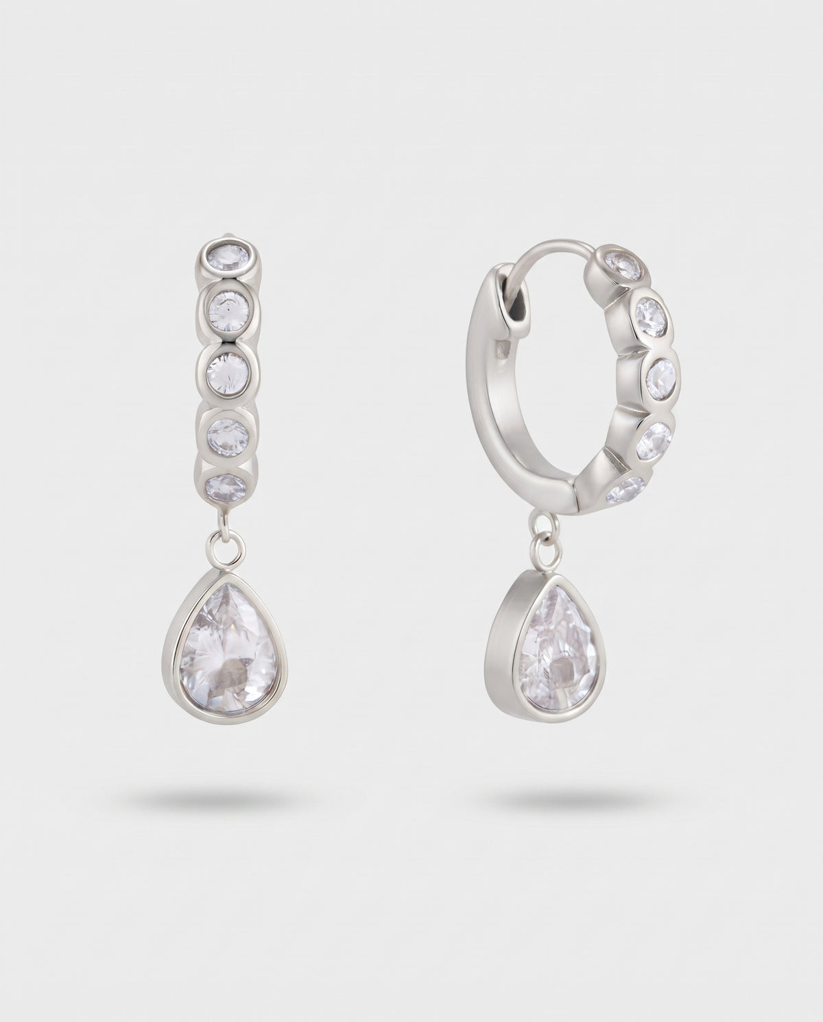 A pair of silver-toned huggie hoop earrings with clear teardrop and round gemstones, displayed on a light gray background.
