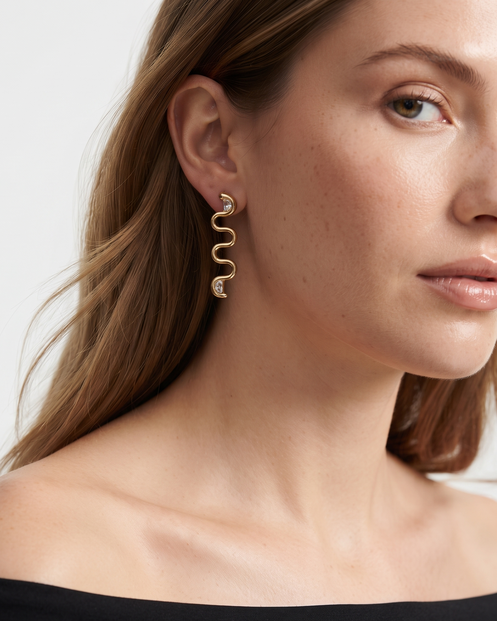 A close-up of a woman wearing a gold wavy drop earring with small clear stones, shown against a light neutral background.