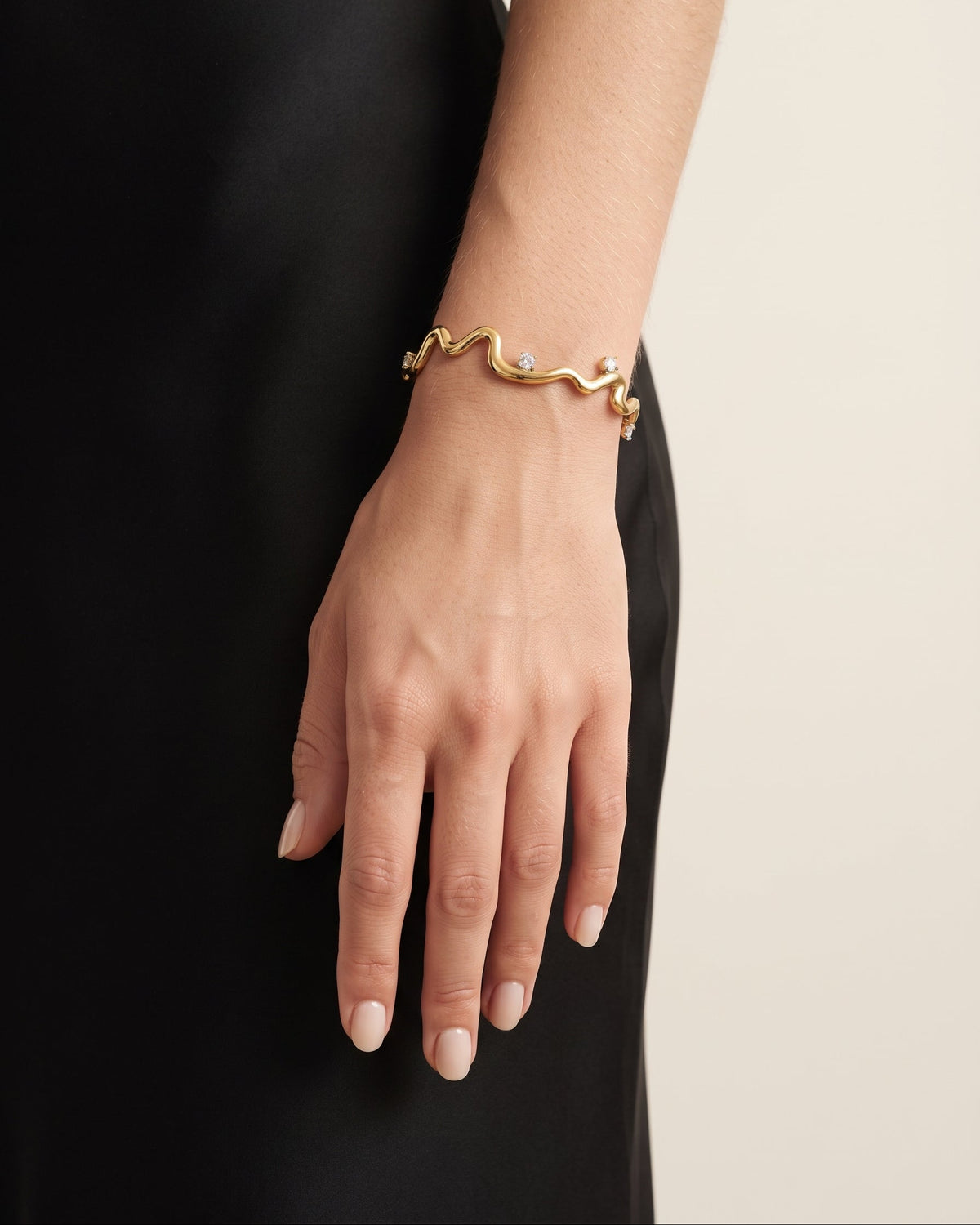 A gold wavy snake-head bracelet with small clear gemstones, worn on a model's wrist against a black dress and a light beige background.