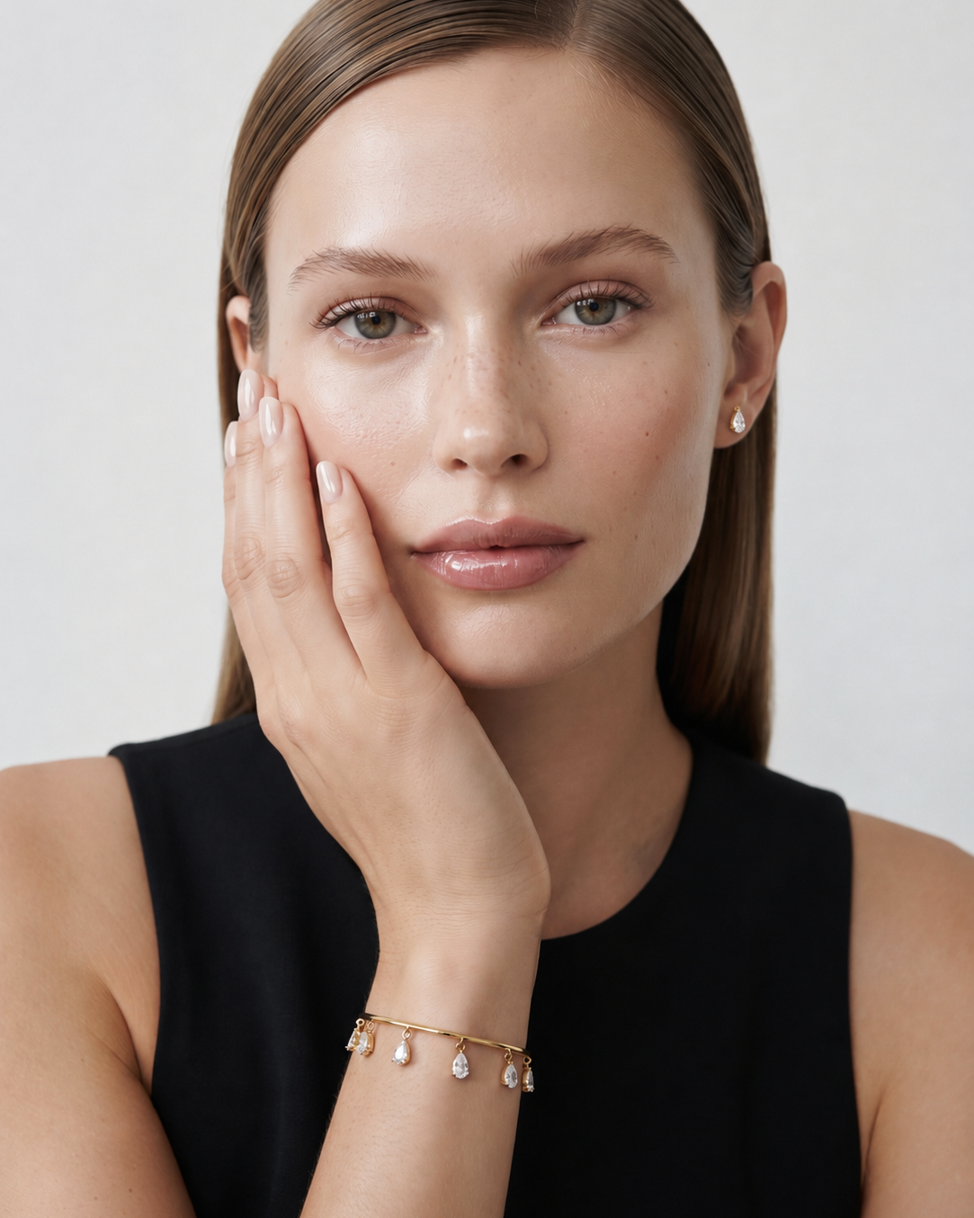 A close-up portrait of a woman with her hand resting on her cheek, wearing a delicate gold chain bracelet adorned with small clear teardrop crystals. The background is a clean, neutral light gray studio backdrop.