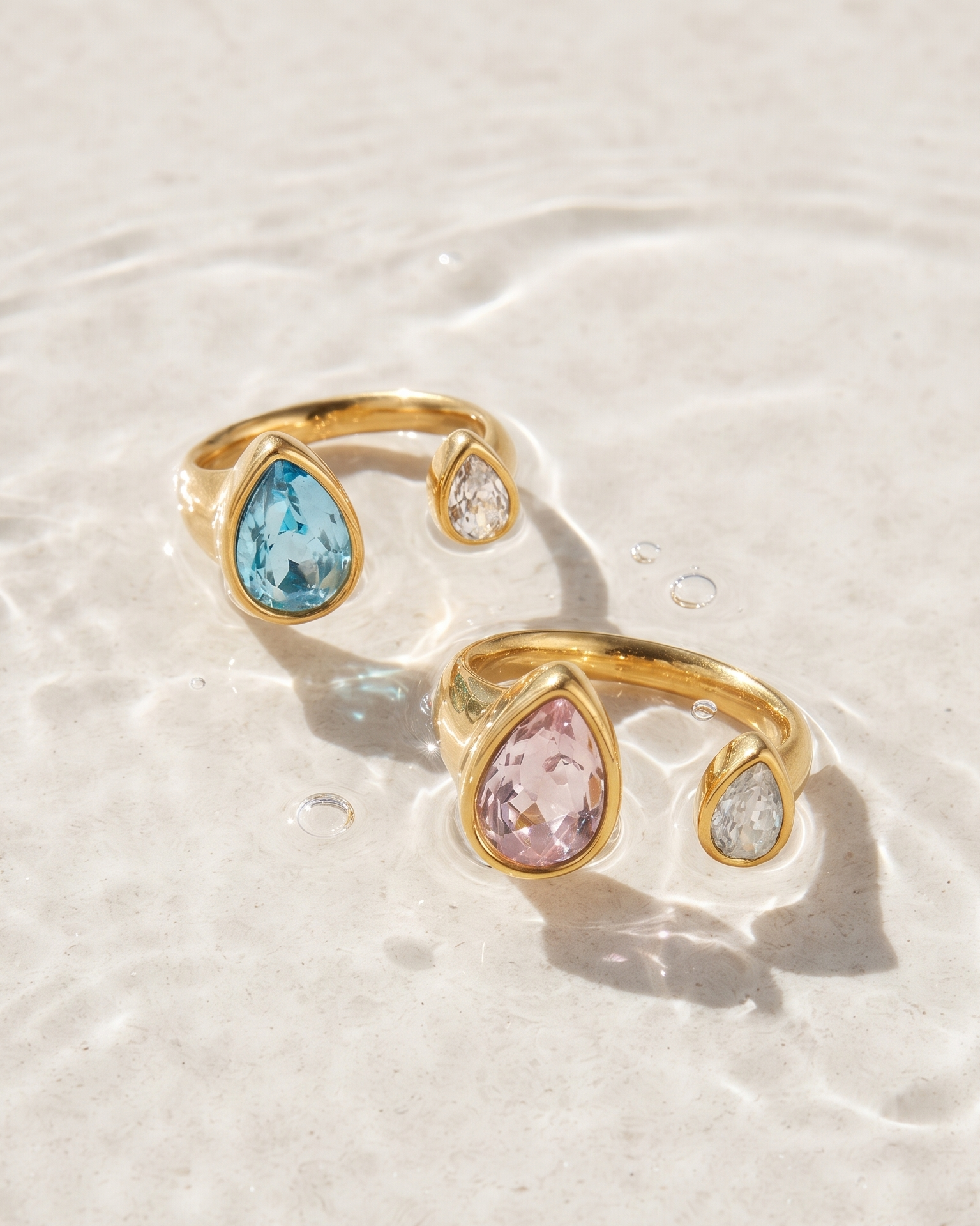 Two gold open bangle rings with teardrop-shaped gemstones, one light blue and one light pink, resting on a surface of rippling clear water.