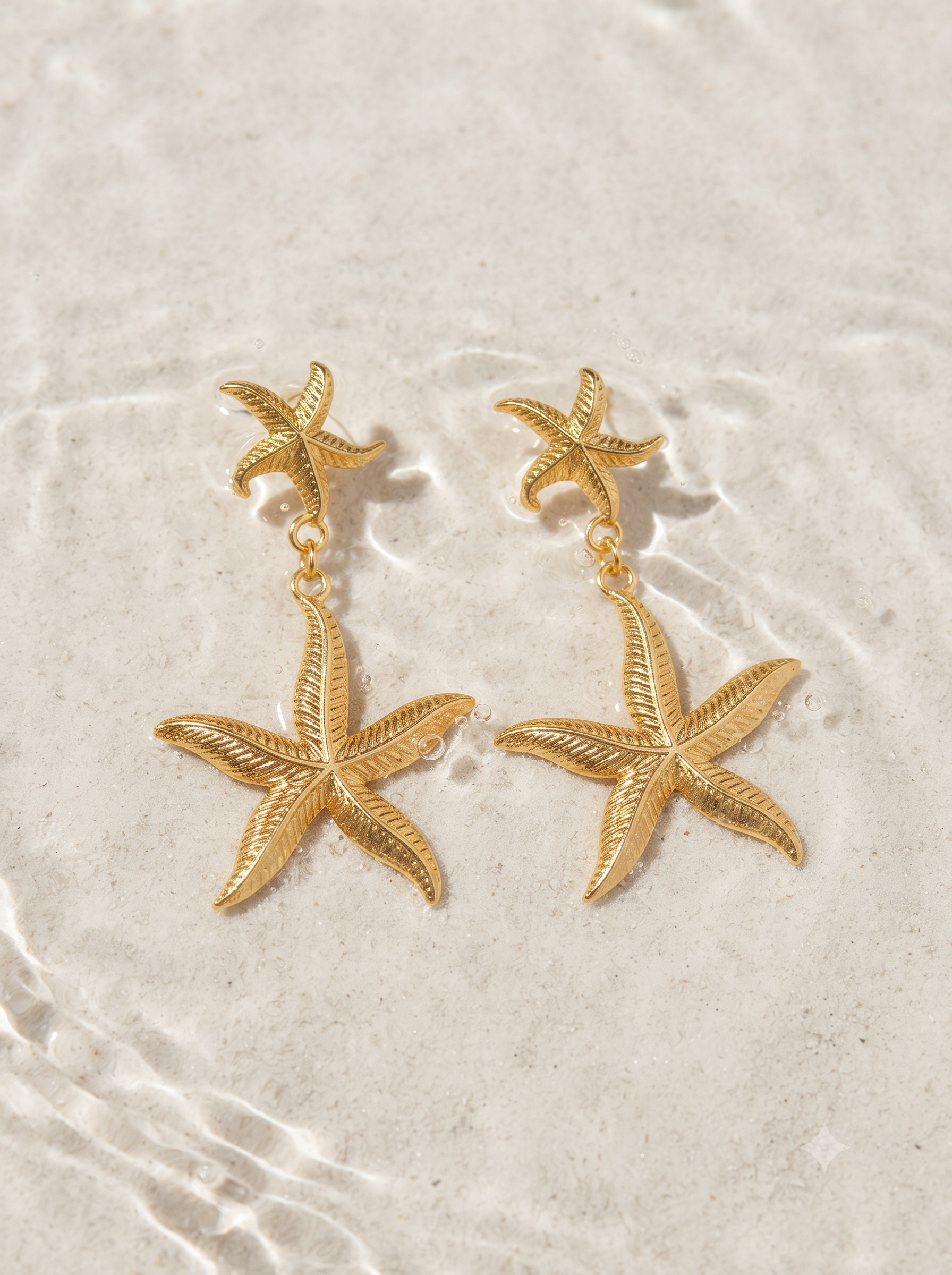 A pair of gold starfish drop earrings displayed on a textured, light beige sandy surface.