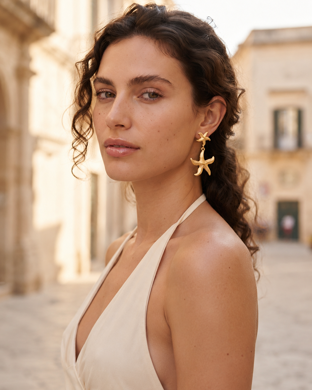 A woman wearing gold starfish-shaped dangle earrings and a cream halter top, photographed outdoors in a sunlit street with blurred buildings in the background.