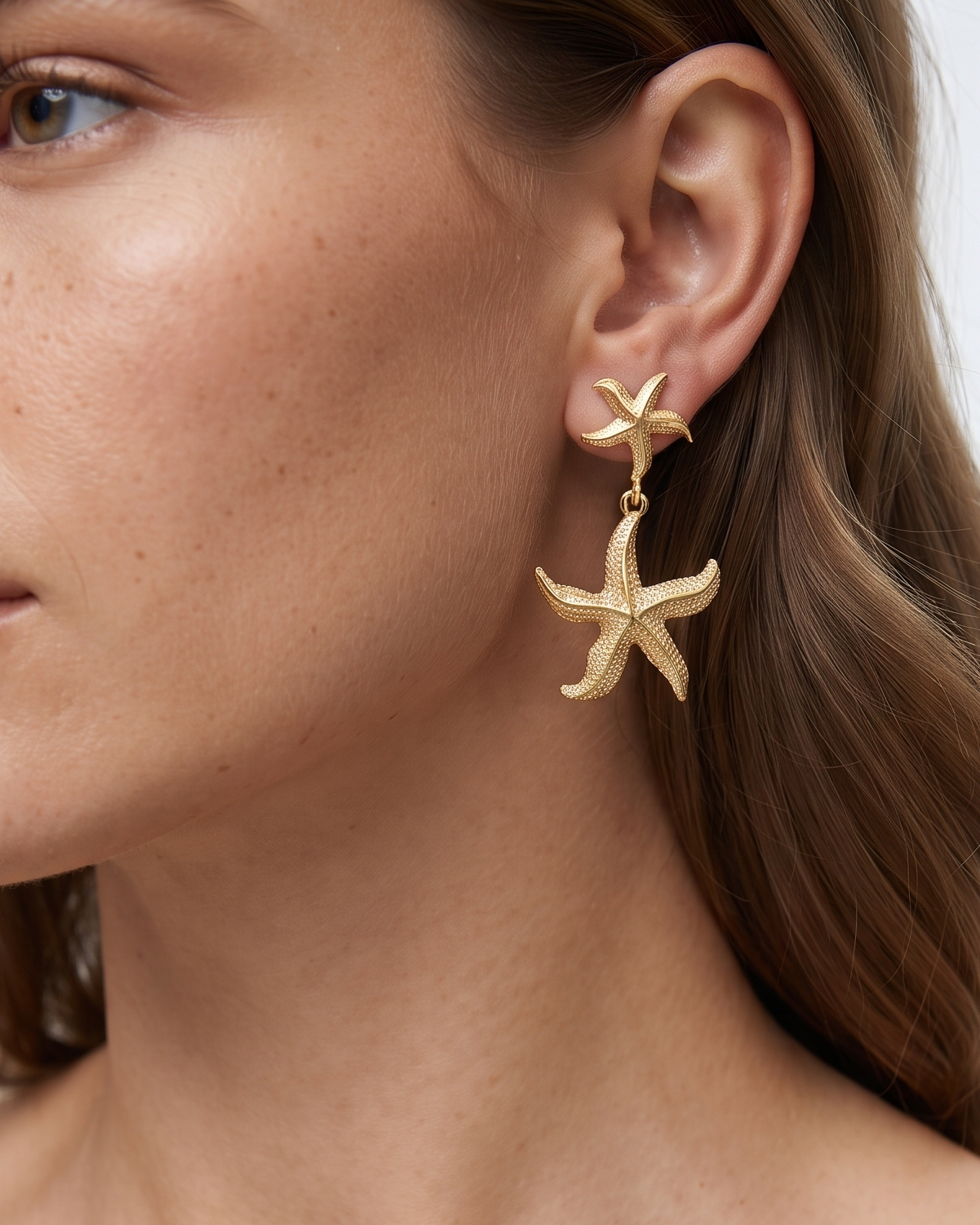 A close-up of a woman wearing a pair of gold starfish drop earrings, showing the jewelry on her ear against her skin and hair.