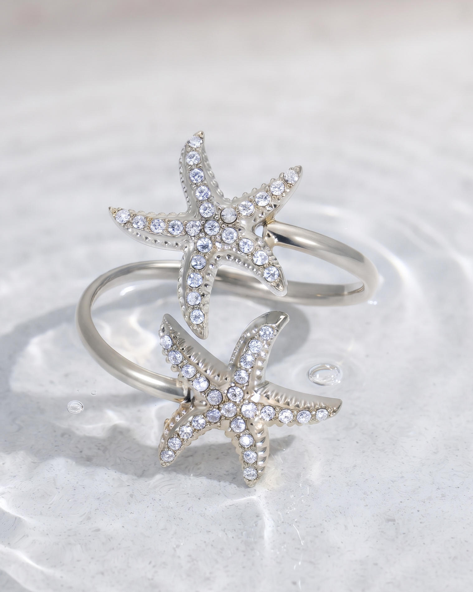 A pair of silver-toned rings featuring starfish designs set with clear rhinestones, resting on a textured, light gray surface with water droplets.