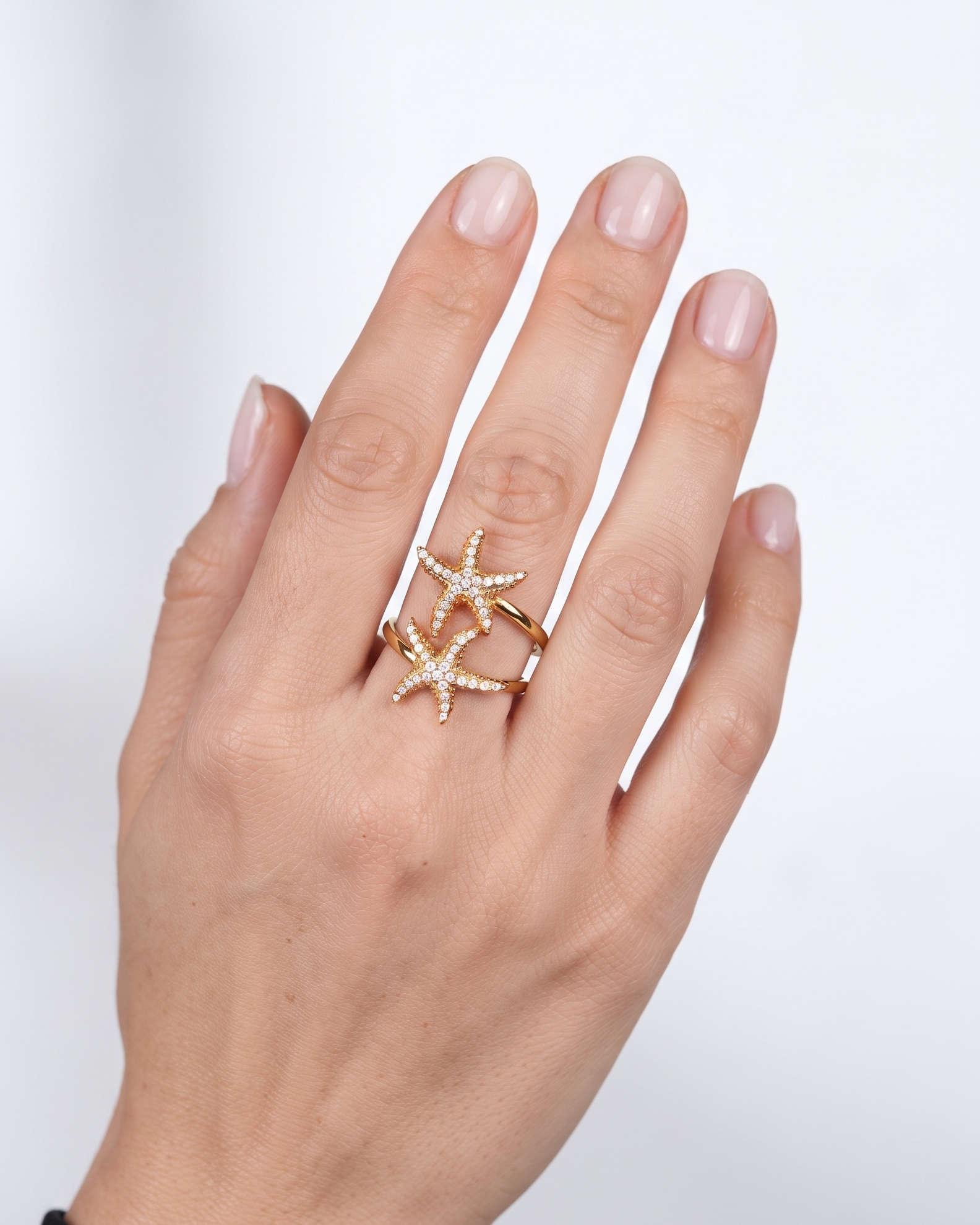 A close-up of a hand wearing a gold ring with two interlocking starfish designs, each adorned with small clear stones, set against a soft light gray background.