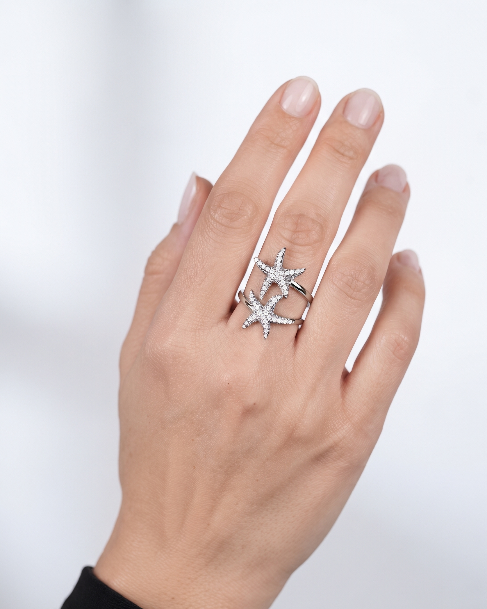 A close-up of a hand wearing a silver ring featuring two interlocking starfish designs set with small clear stones, against a soft light gray background.