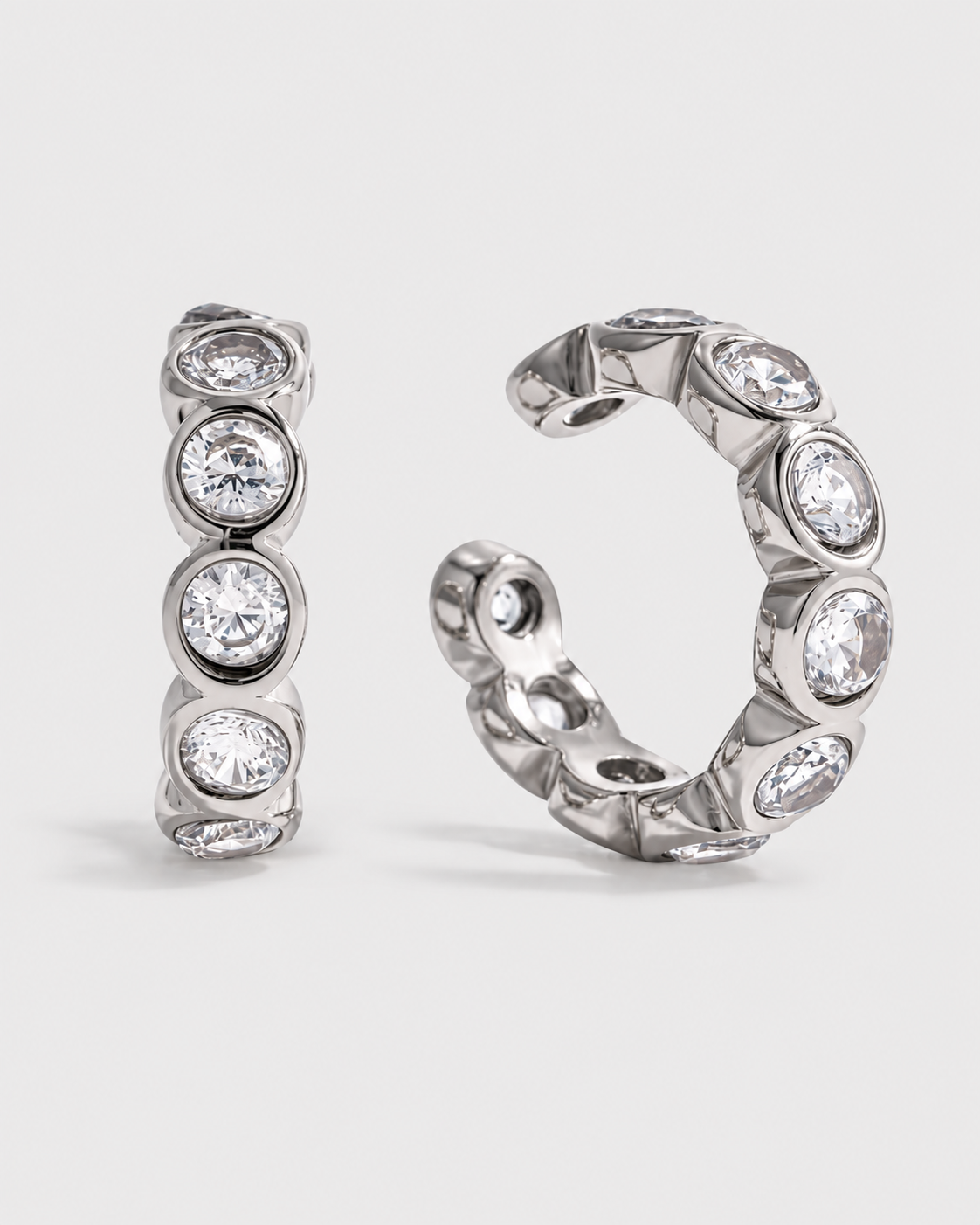 A pair of silver huggie hoop earrings with round clear gemstones, shown on a light gray background.