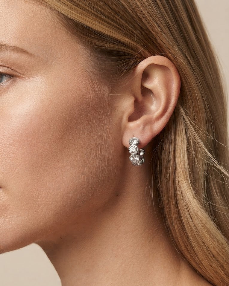 A close-up of a model's ear wearing a small silver huggie hoop earring adorned with clear gemstones.