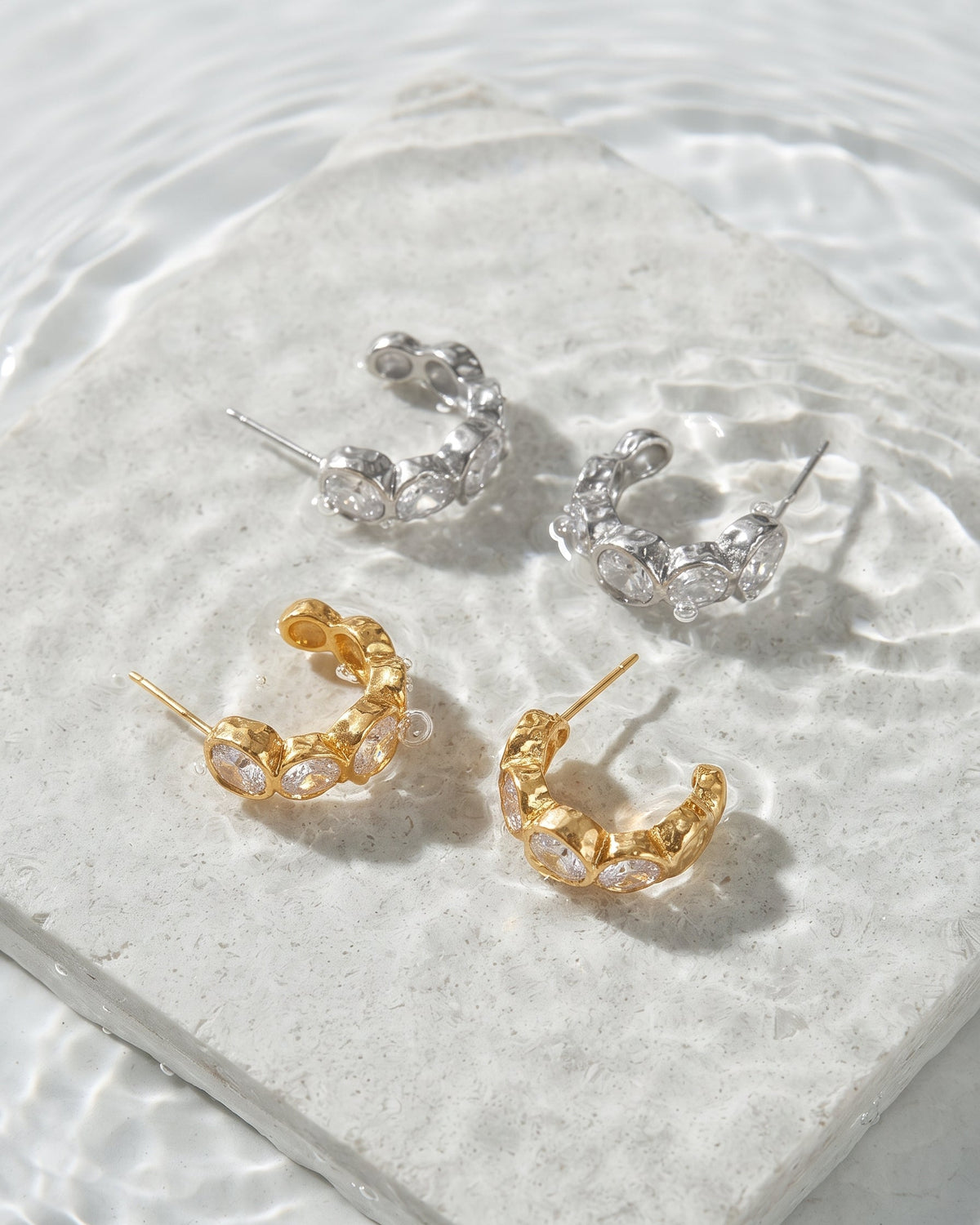 Four small hoop earrings arranged on a light stone slab with rippling water around it. Two hoops are silver-toned with clear faceted stones, and two are gold-toned with clear faceted stones.