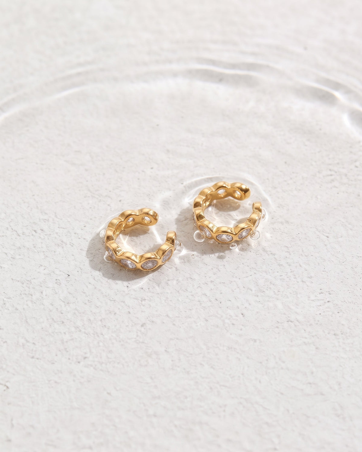 A pair of small gold huggie hoop earrings with clear oval gemstones, resting on a textured white surface with a shallow pool of water around them.