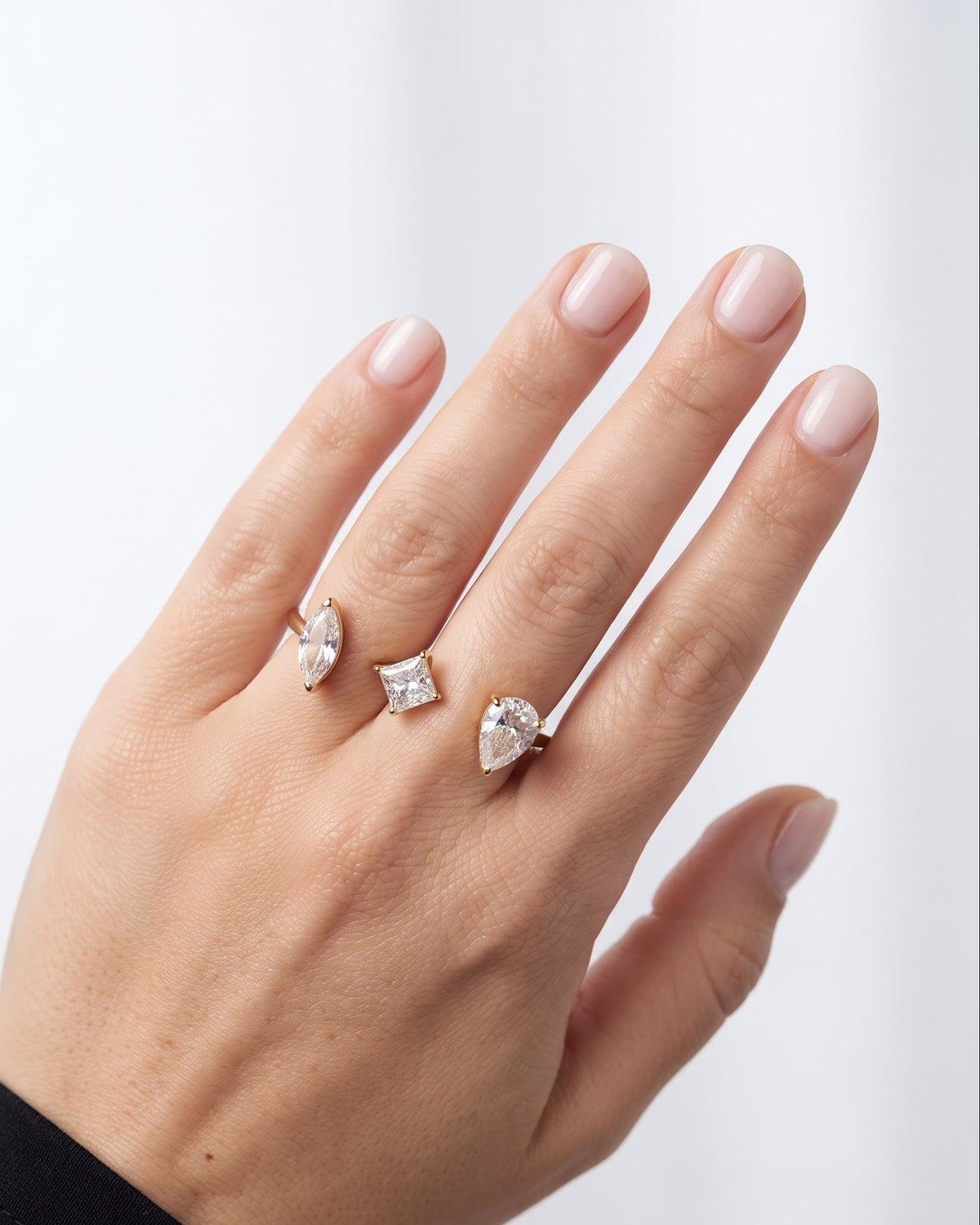 A close-up of a hand wearing three silver rings with clear gemstones in different shapes, set against a soft, light gray background.
