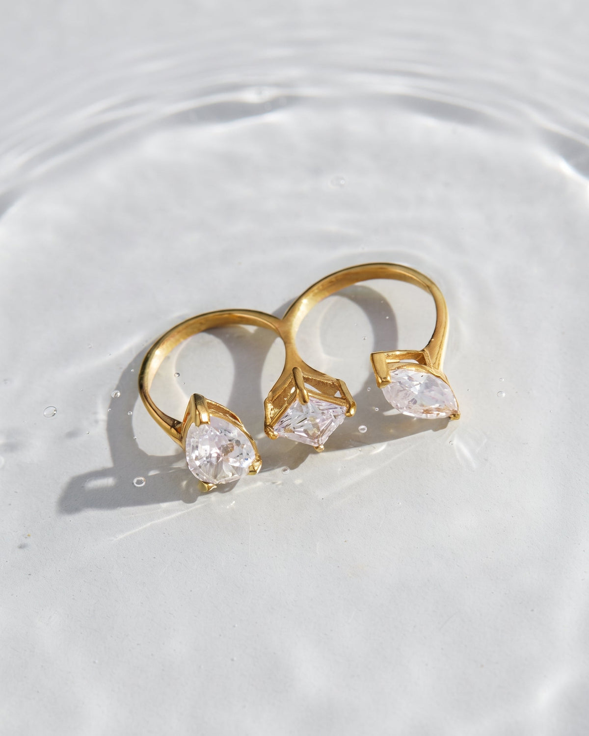 Two gold rings with marquise-cut clear gemstones, resting on a white surface with soft water ripples and shadows.
