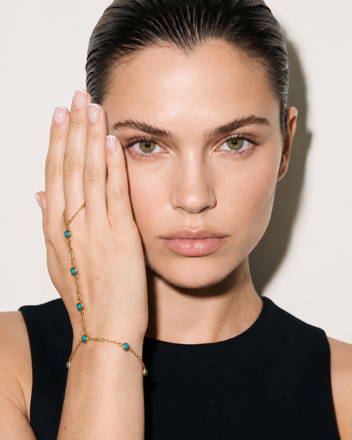 A close-up of a model wearing a delicate gold chain bracelet and matching ring with small turquoise blue beads, posing with her hand against a clean off-white background.