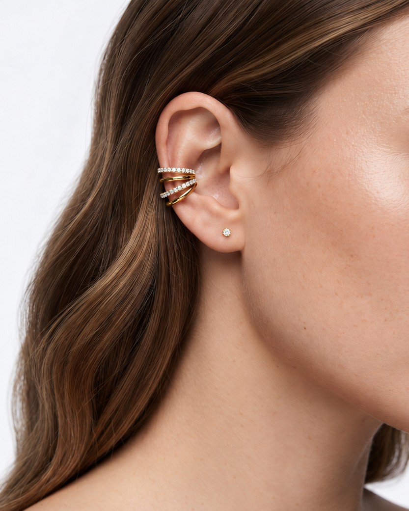 A close-up of a woman's ear wearing a gold triple-hoop ear cuff with small clear stones, alongside a tiny round stud earring, against a clean white background.