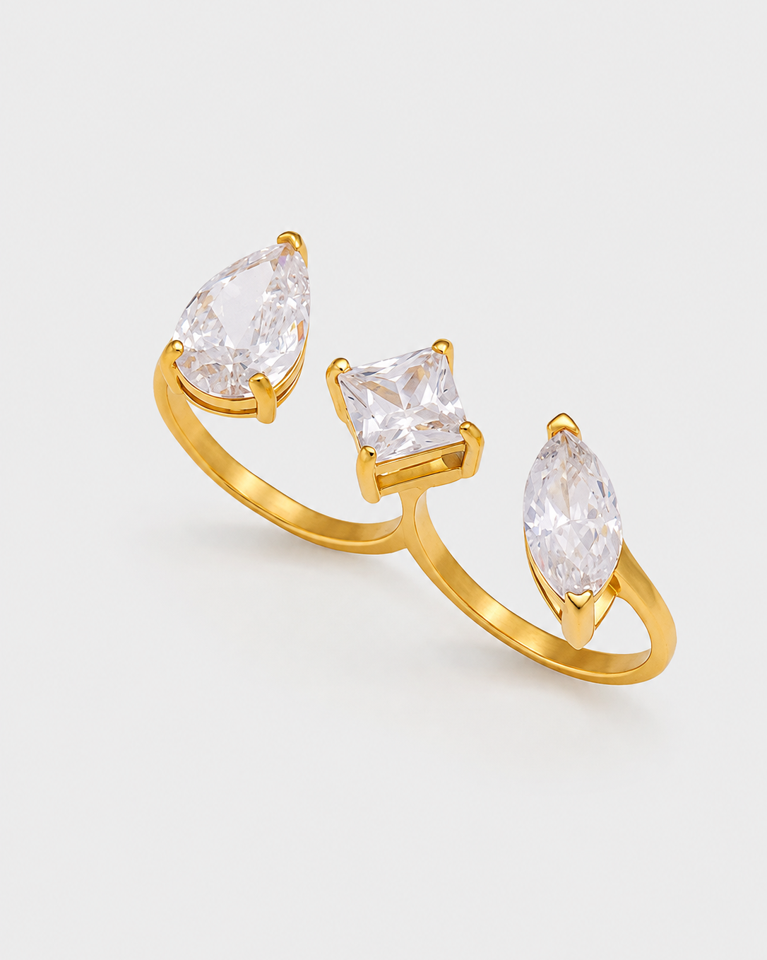 A gold ring featuring three clear gemstones in pear, square, and marquise cuts, displayed on a light gray background.