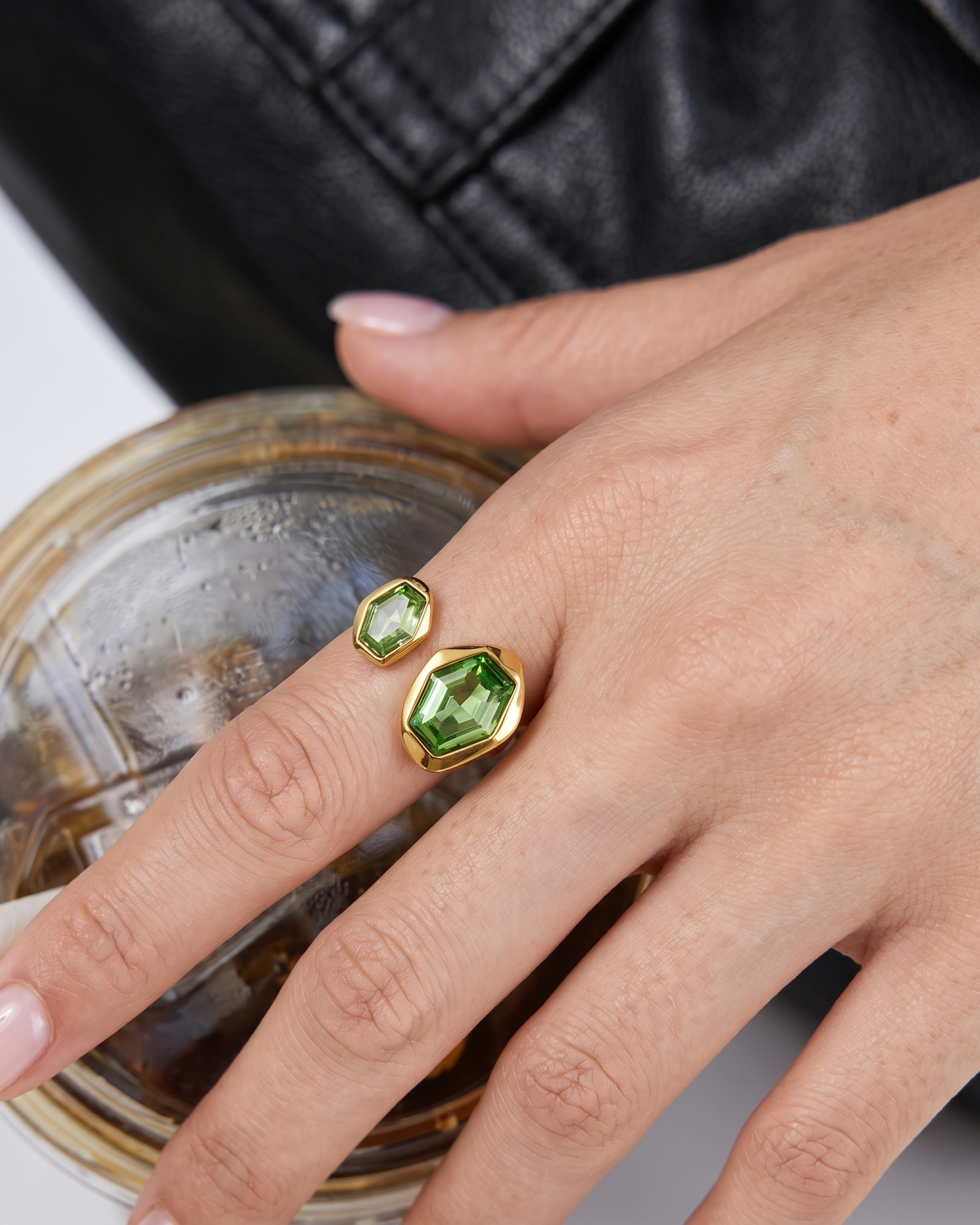Hand wearing a gold ring with green gemstones, holding a drink.