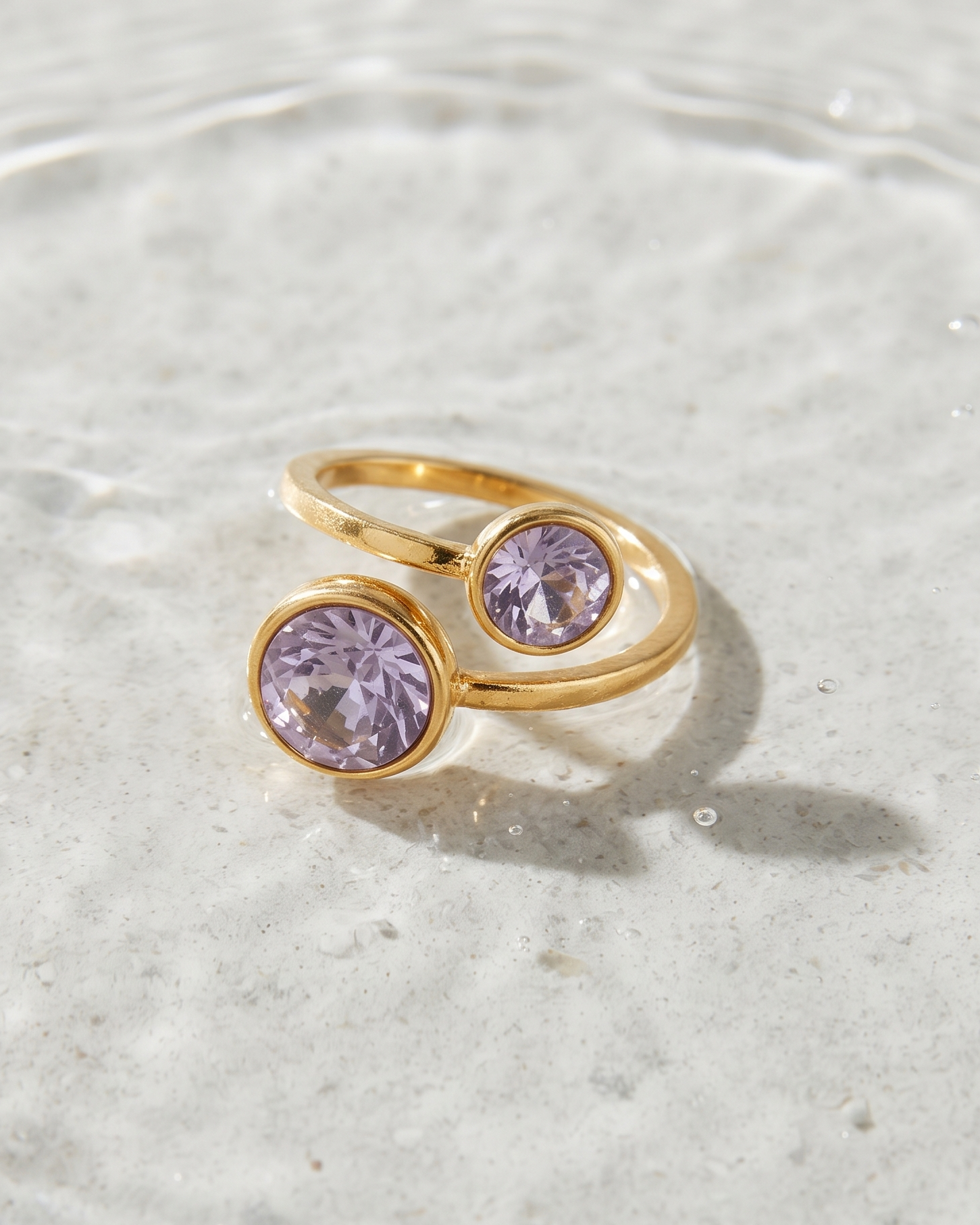 A gold ring with two round light purple gemstones, resting on a textured light gray stone surface with shallow water ripples in the background.