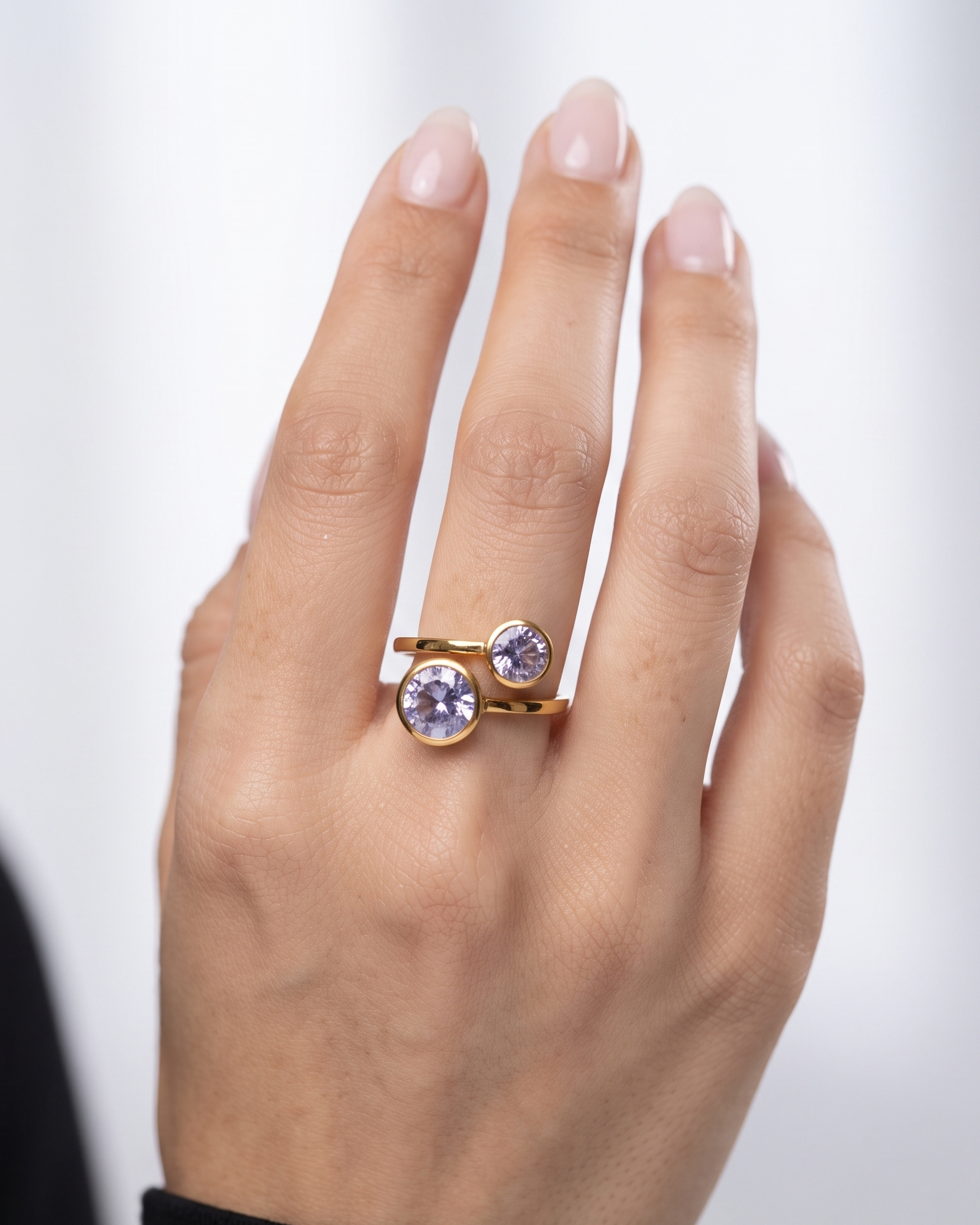 A close-up of a hand wearing a gold ring with two round bezel-set light purple gemstones, set against a soft, light gray background.