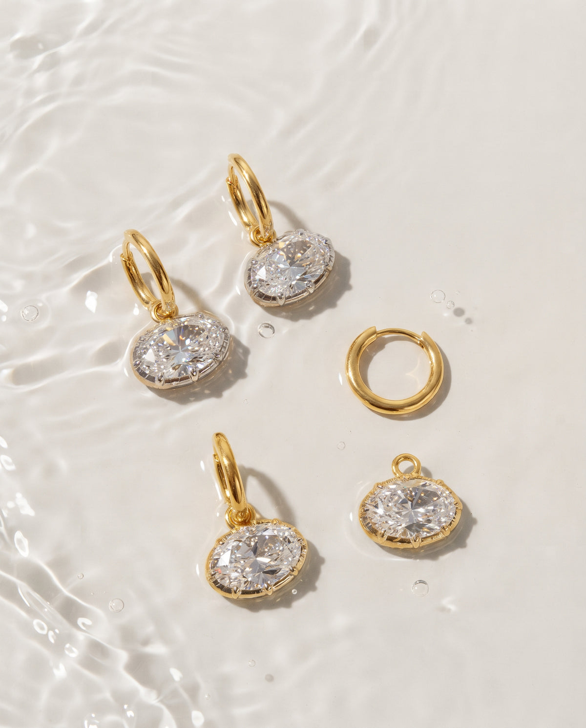 Gold jewelry pieces including two drop earrings, a matching hoop earring, and a small charm, arranged on a glossy white surface with water ripples.