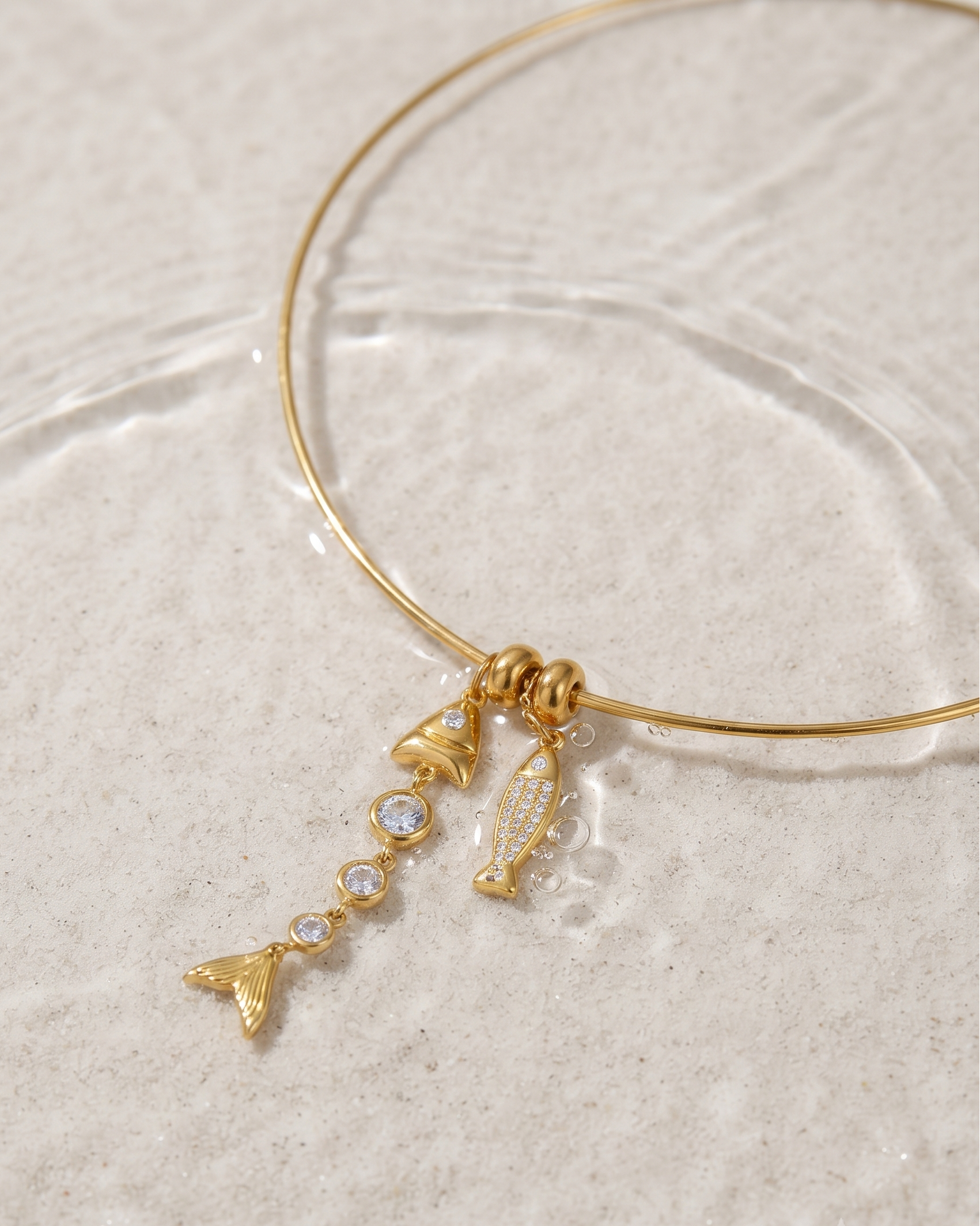 A delicate gold wire bangle bracelet with dangling charms, including a small mermaid tail, a shell, and a pearl-like bead, displayed on a textured beige surface with water ripples.