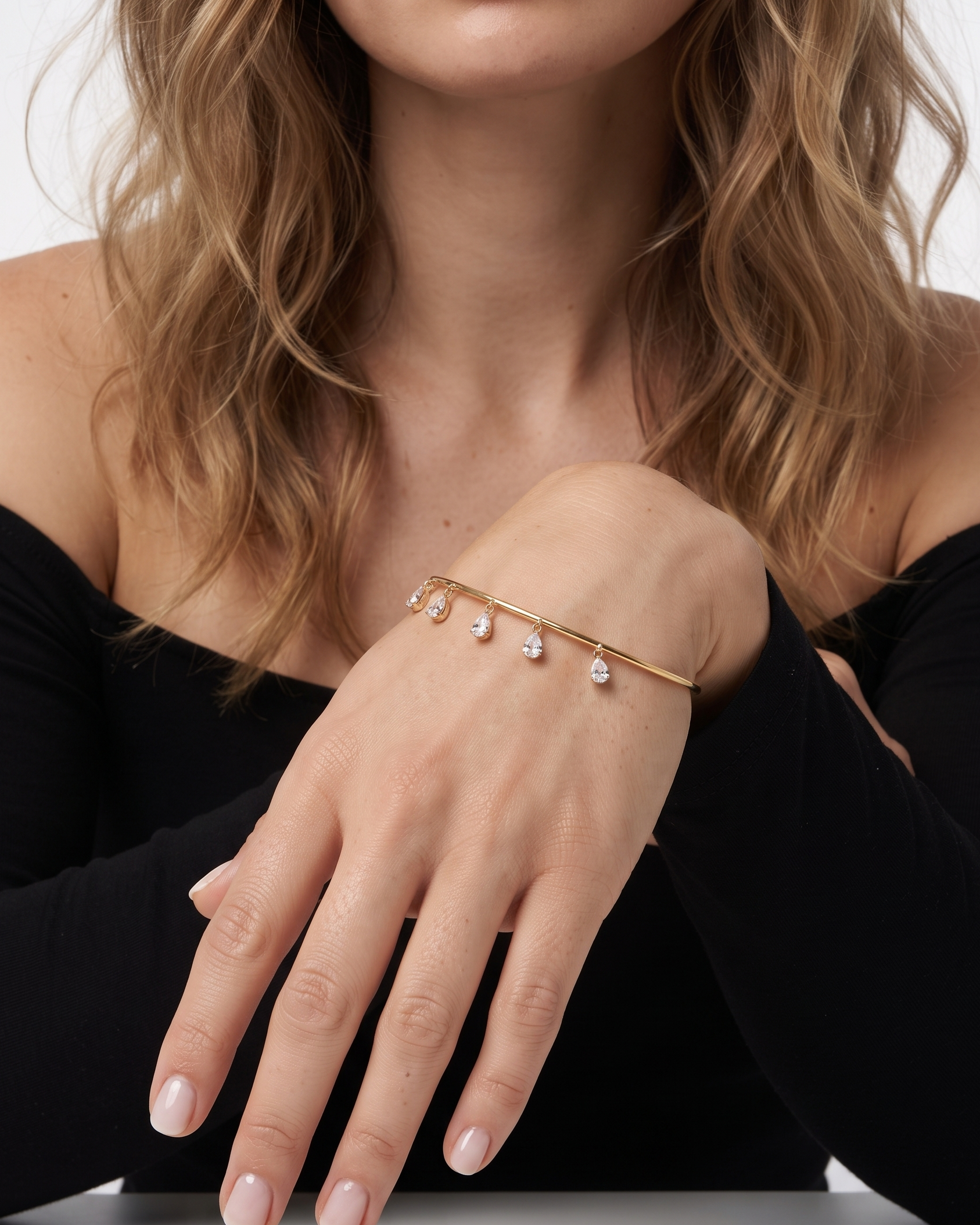 A close-up of a model's hand wearing a delicate gold bangle bracelet adorned with small teardrop-shaped clear stones, set against a clean white background.